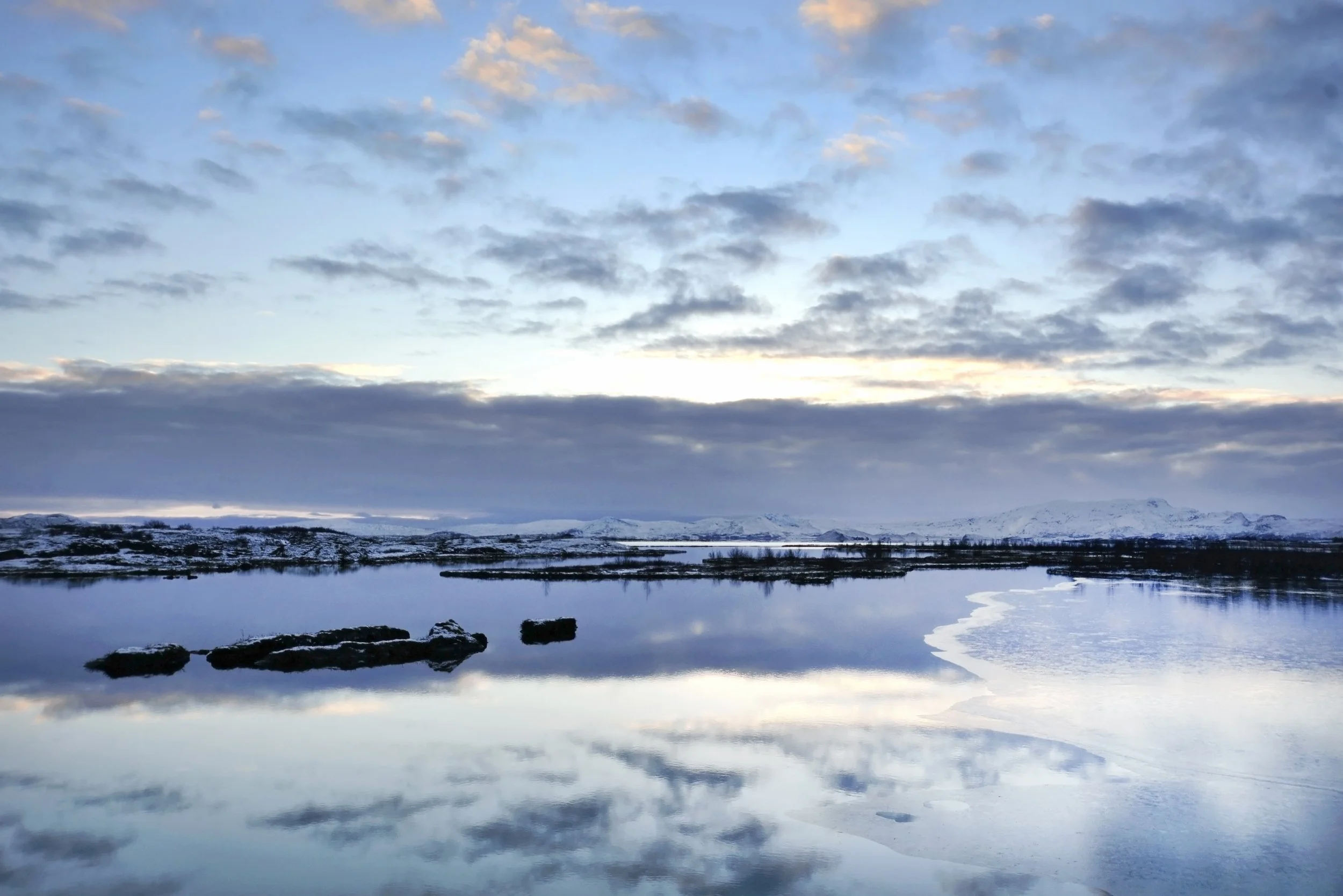 A serene winter landscape showing a calm body of water reflecting the cloudy sky, with snow-covered mountains in the distance.