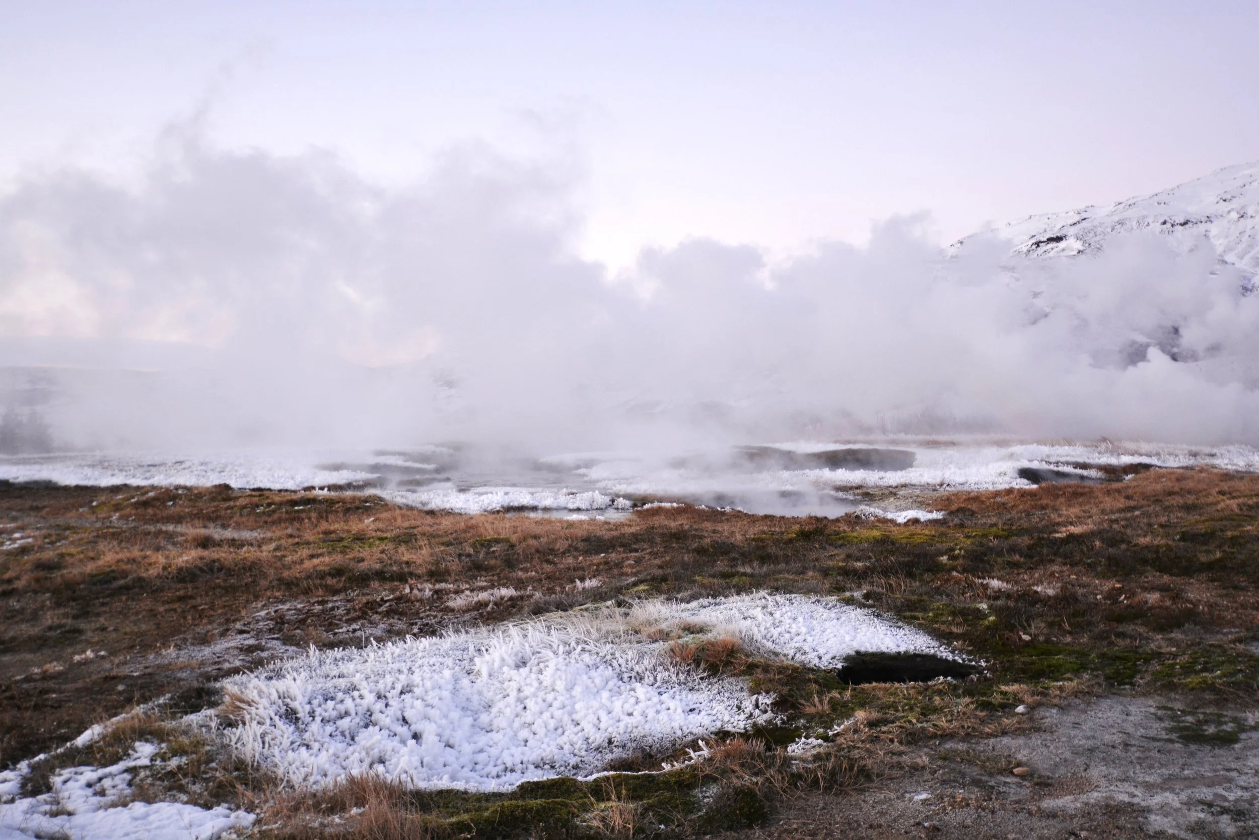 A geothermal area with boiling water and steam rising from hot springs, with snow and ice on the ground and mountains in the background.