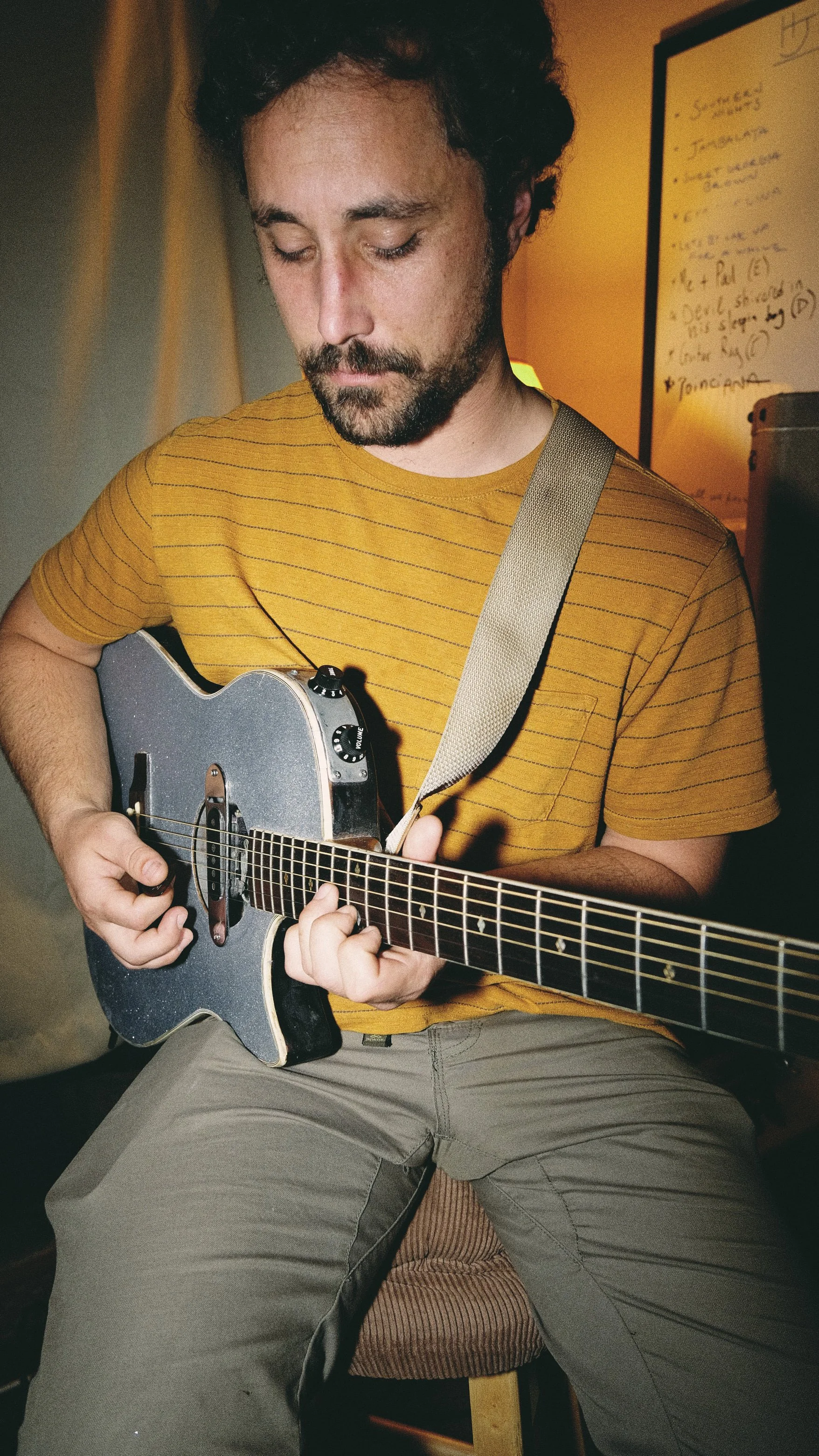 A man with dark curly hair and a beard playing an electric guitar indoors, wearing a yellow striped T-shirt and khaki pants.