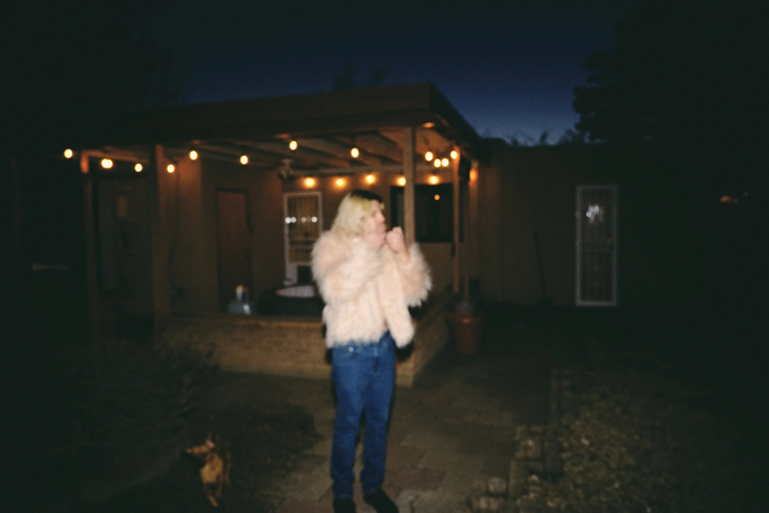 A woman with blonde curly hair wearing a fuzzy beige jacket and blue jeans standing outside at night near a house with string lights.