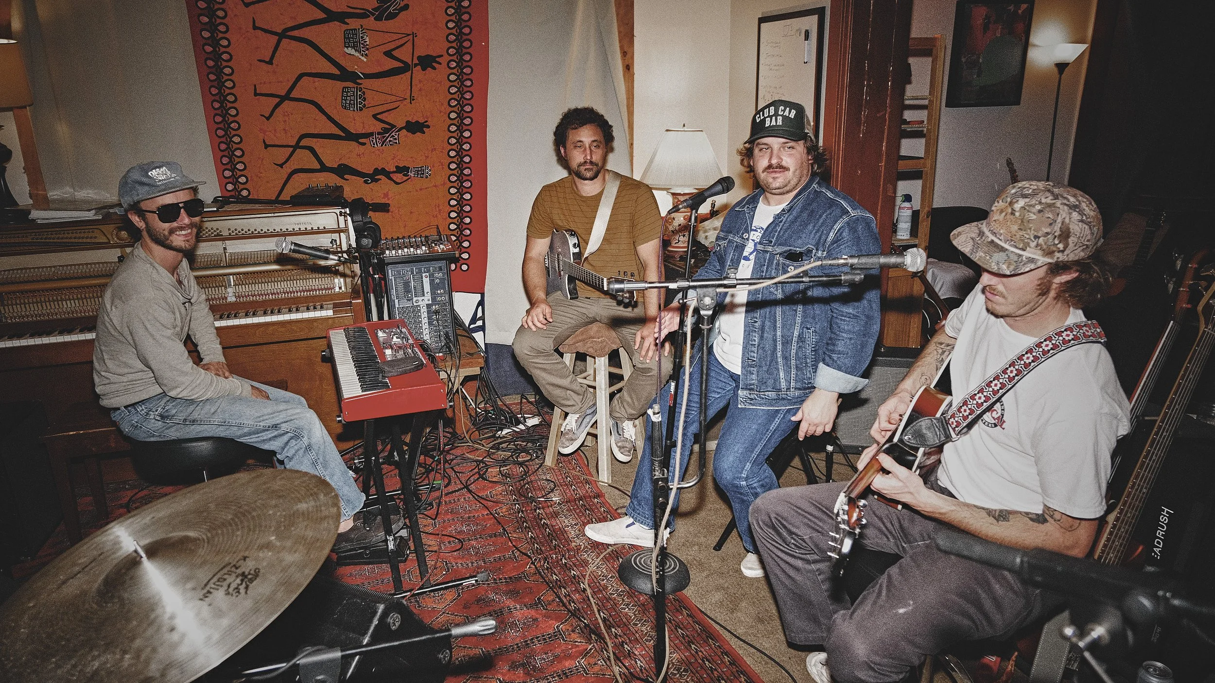 A band performing in a cozy room with musical instruments, including guitars, a keyboard, and drums, and colorful artwork on the wall behind them.