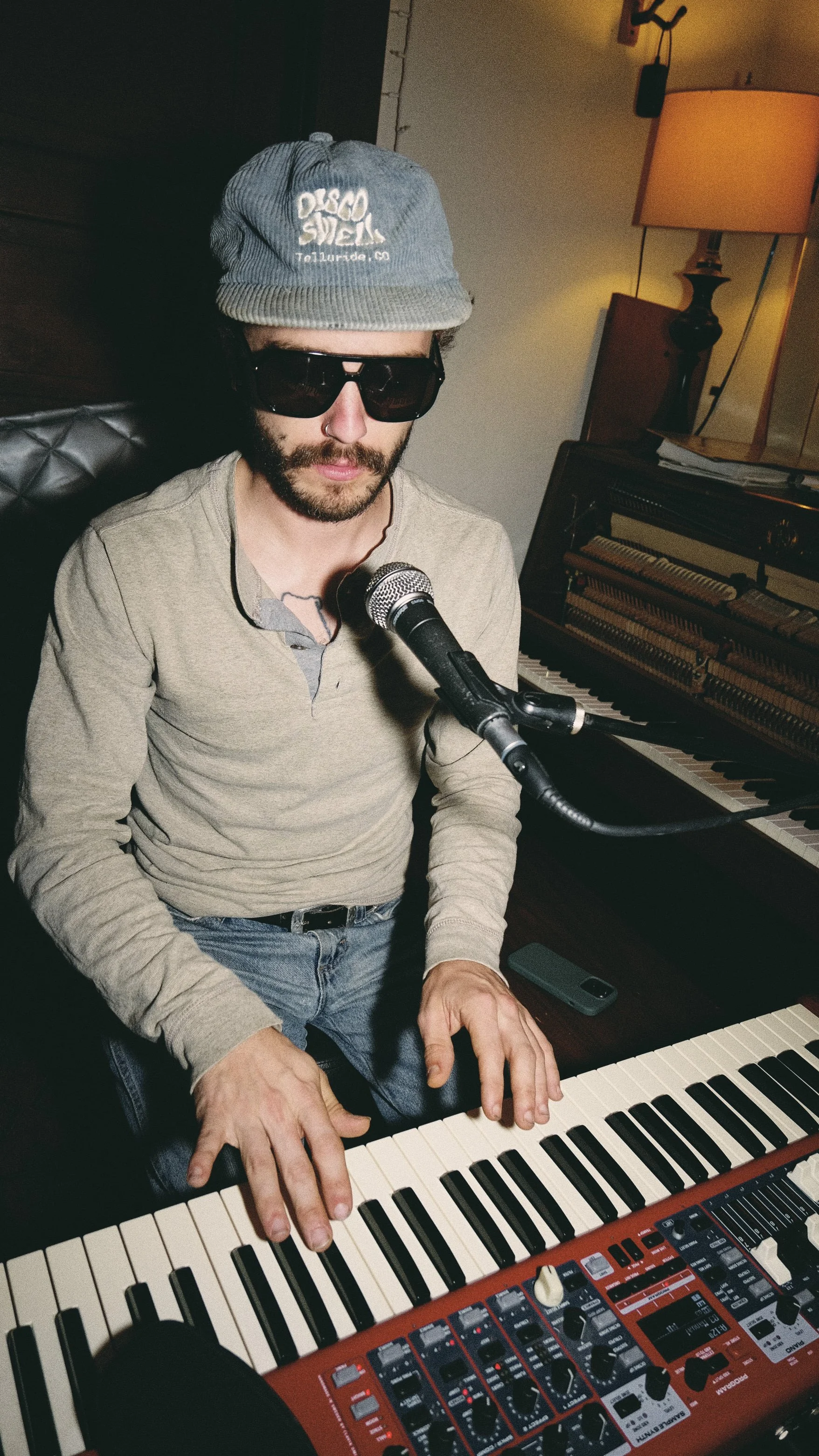 A man wearing sunglasses, a gray beanie, and a beige long-sleeve shirt playing a keyboard with a microphone in front of him in a room with a piano and a lamp.