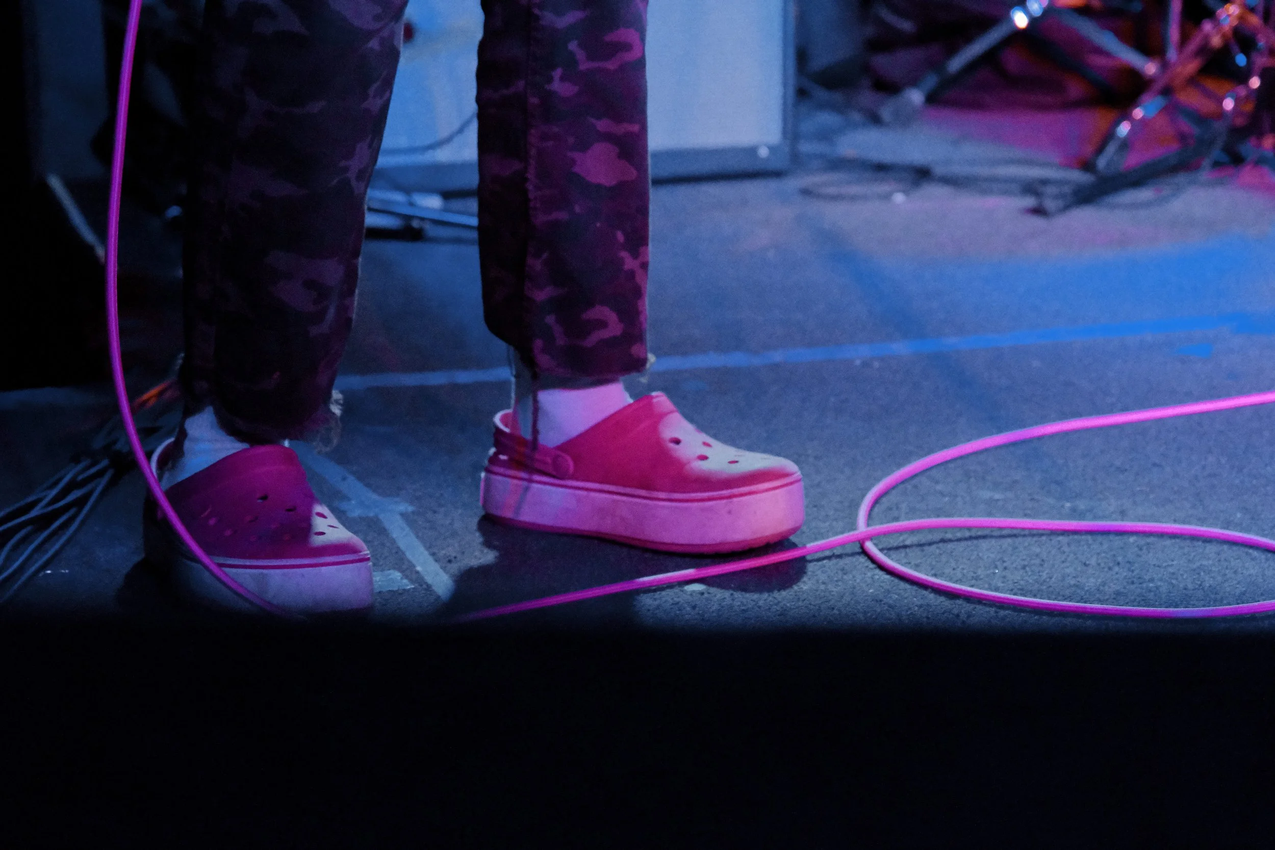 Close-up of a person's feet wearing white platform shoes with pink accents, standing on a dark stage with pink cables and equipment in the background.