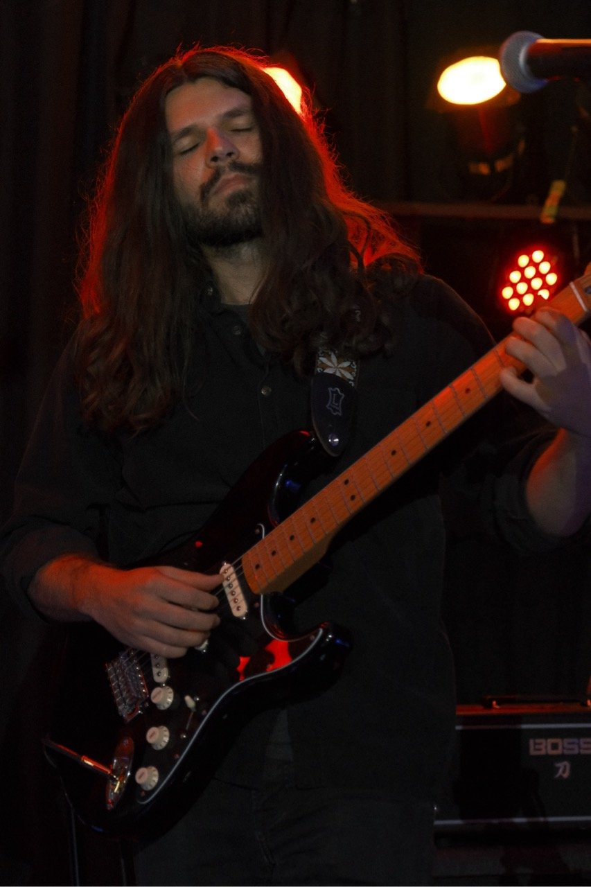 A man with long brown hair and a beard is playing an electric guitar on stage with closed eyes and a calm expression, illuminated by warm stage lights.