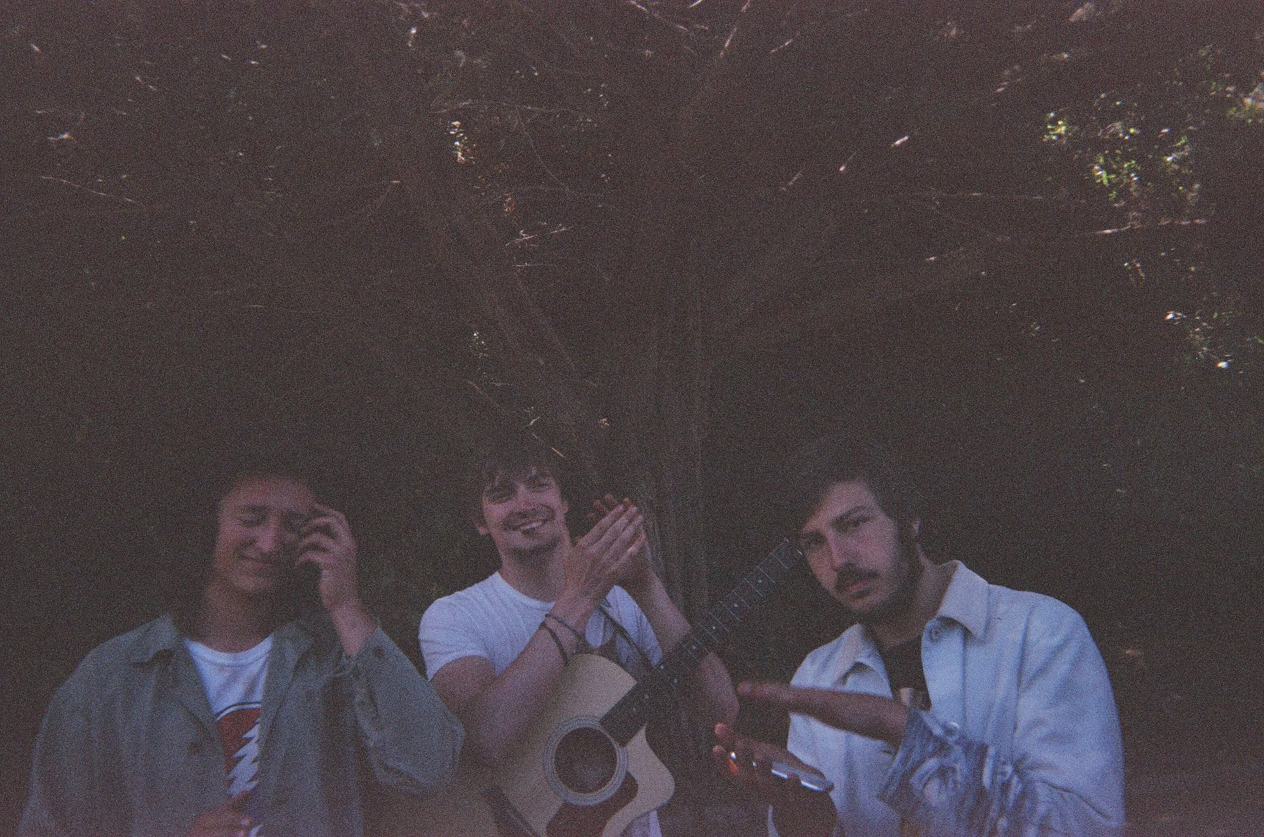 Three young men outdoors at night, one holding a guitar, one with a phone to his ear, and the third pointing and making a gesture, under a dark tree canopy.