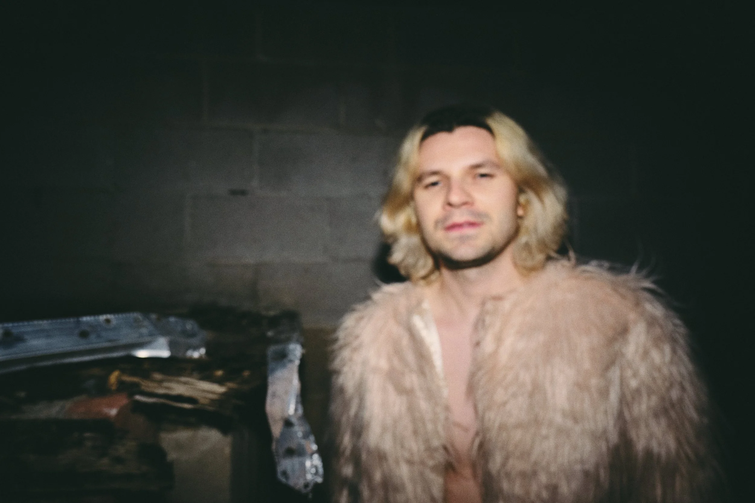 A man with shoulder-length blonde hair and a light beard wearing a tan fur coat standing indoors against a dark wall.