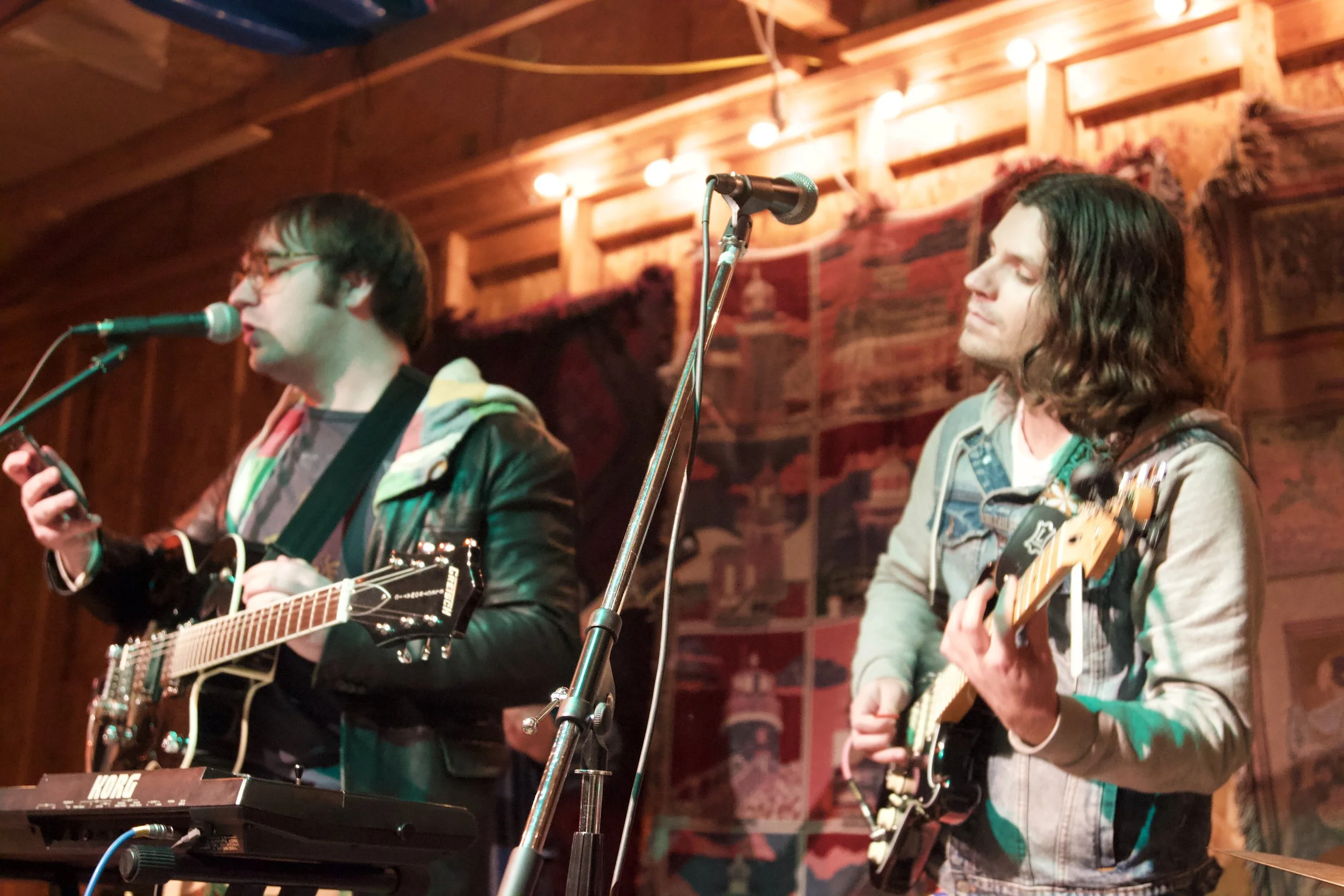 Two men perform on stage with guitars, microphones, and a small electronic keyboard, in a cozy, wood-paneled venue.