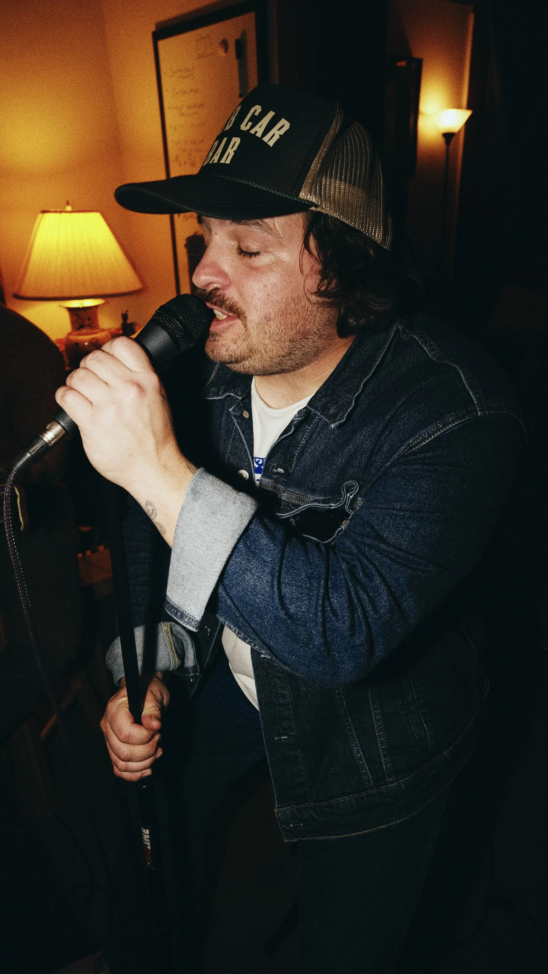 A man singing into a microphone in a dimly lit room, wearing a trucker hat and denim jacket.