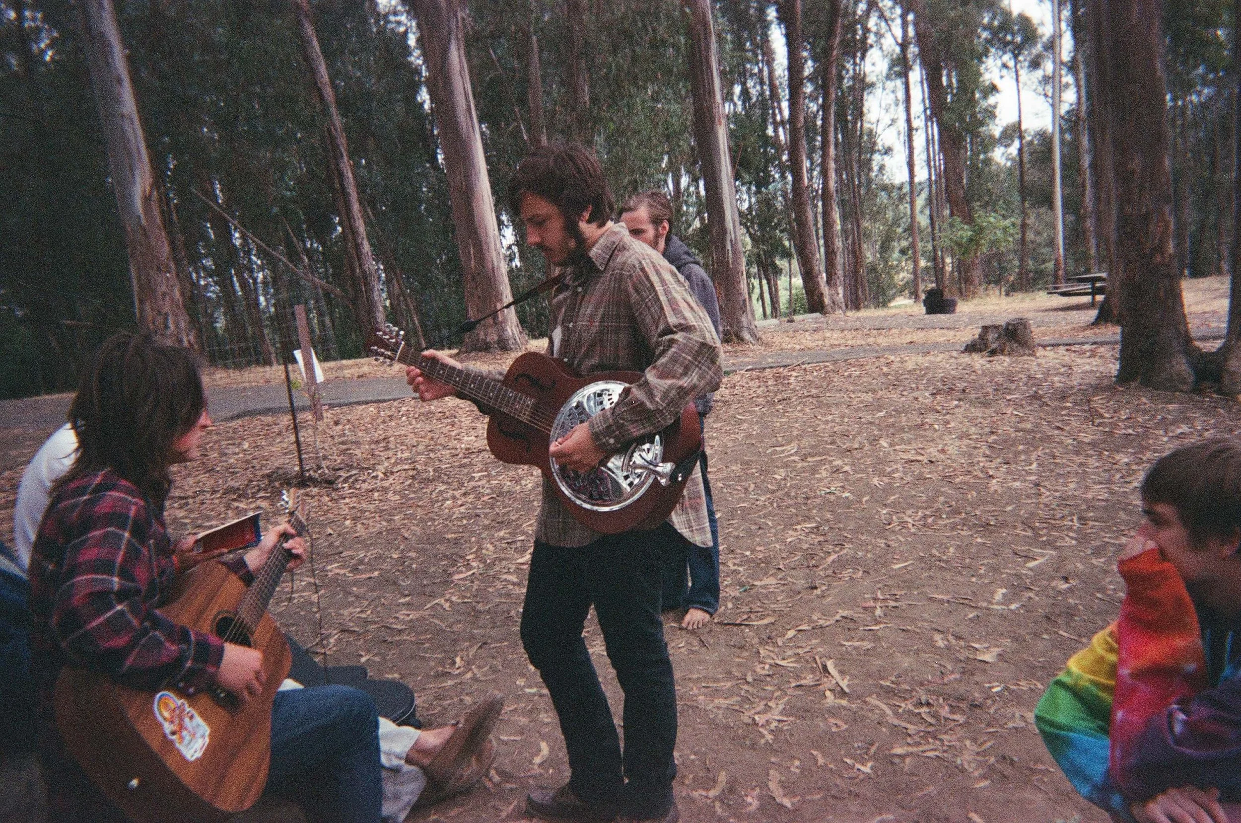 Group of young people playing guitars in a wooded park area, some sitting and one standing, during daytime.