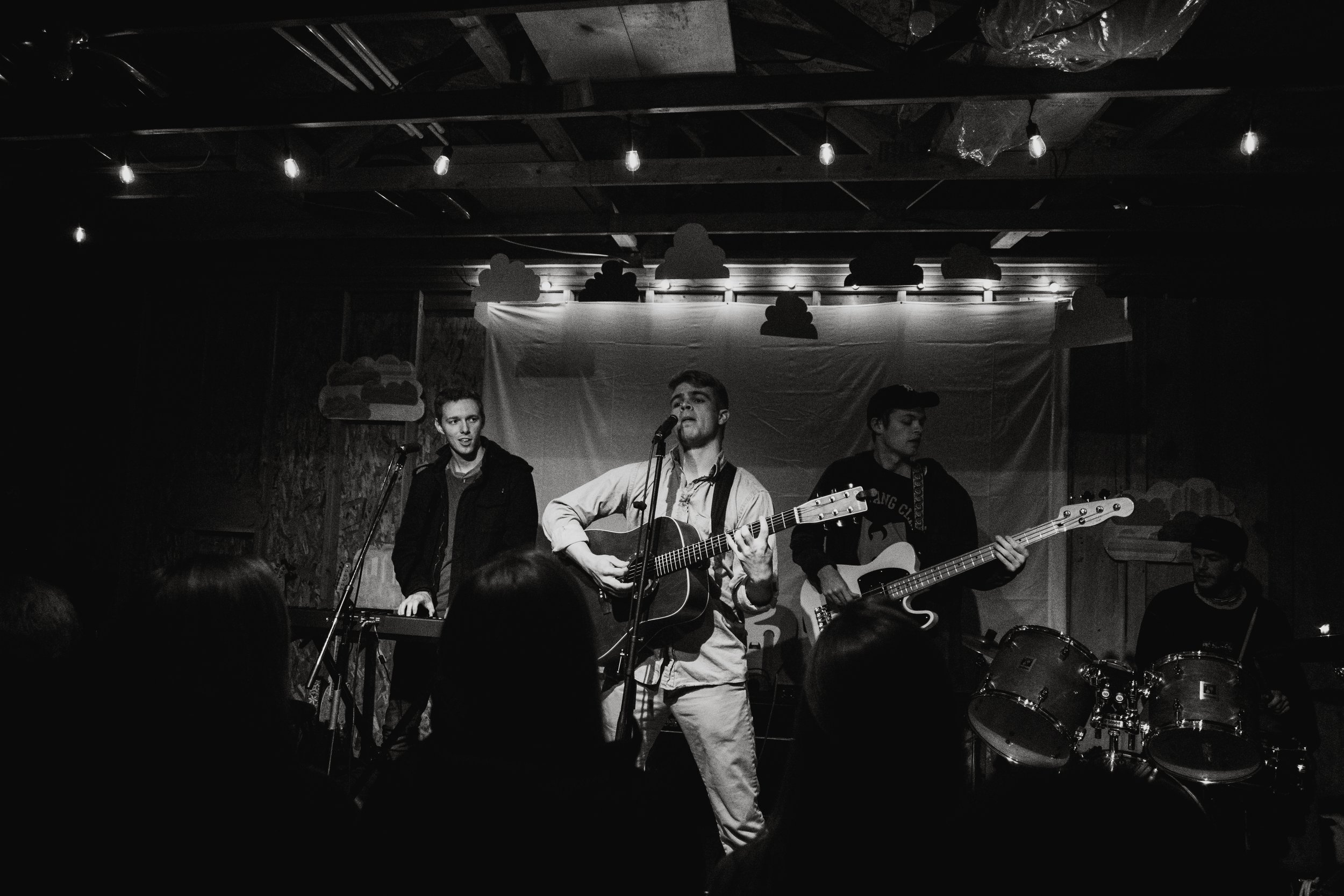 A black and white photo of a four-member band performing on stage. The lead singer is singing and playing an acoustic guitar, with two other band members playing electric guitar and drums. There is a simple backdrop, and the audience is visible in th