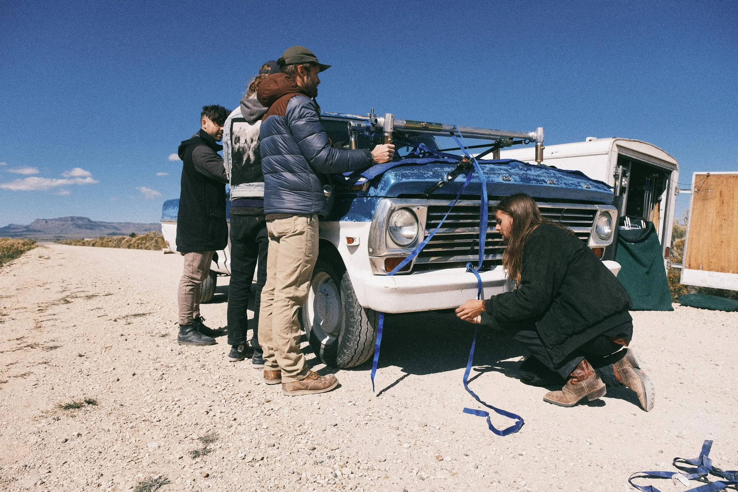 Group of people working together to secure a blue van with straps in a desert landscape under a clear blue sky.