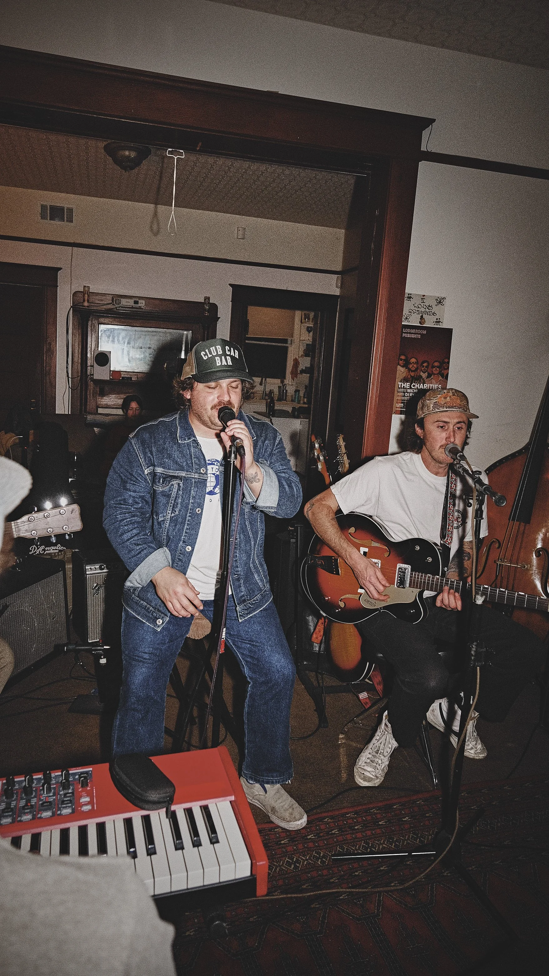 Two musicians performing at a small indoor venue. One is singing into a microphone and the other is playing an electric guitar. The man on the left wears a denim jacket and a cap, while the man on the right wears a white t-shirt and a camouflage cap.