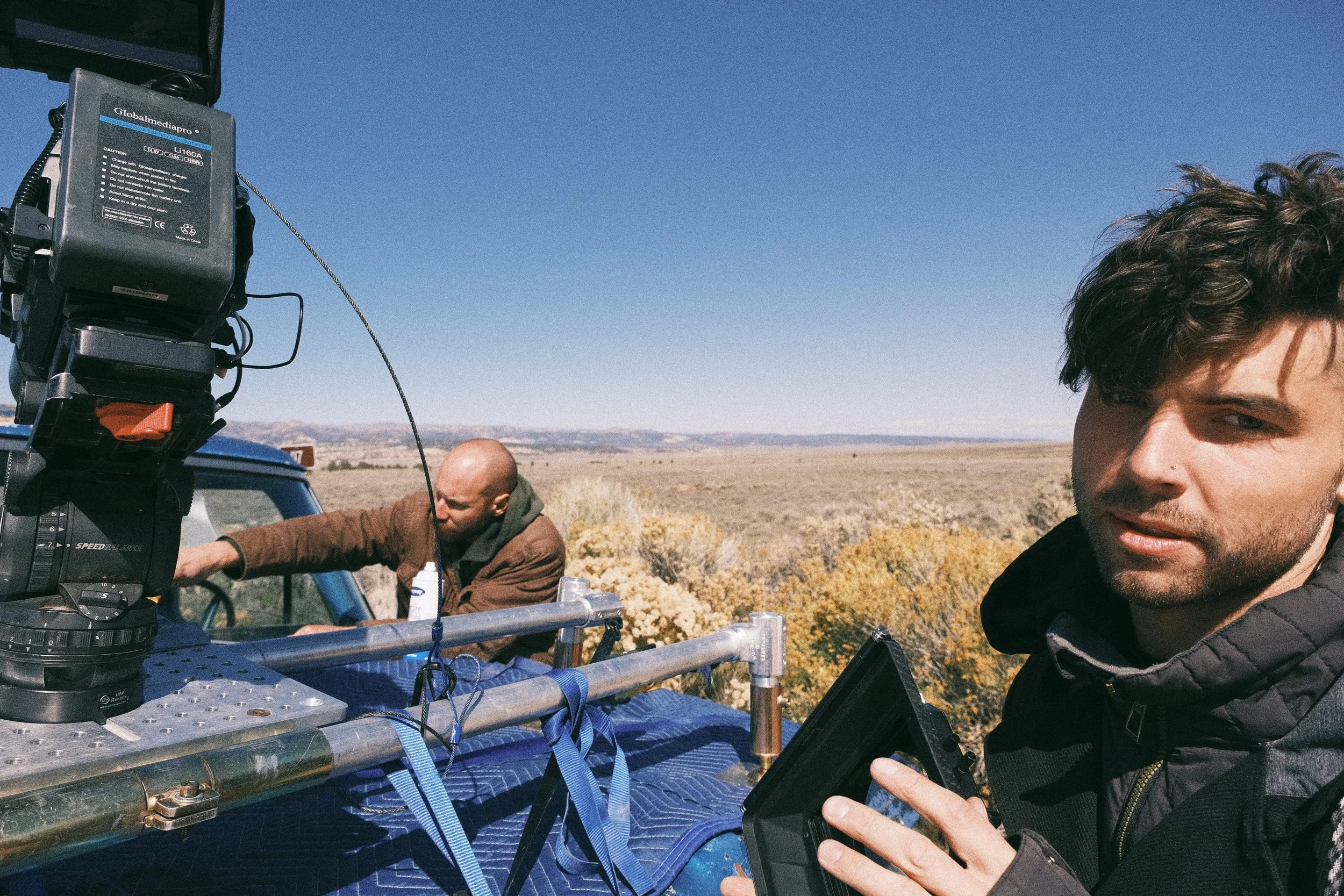 Two men working outdoors with filming equipment on a vehicle, set against a wide, open landscape with desert vegetation and a clear blue sky.