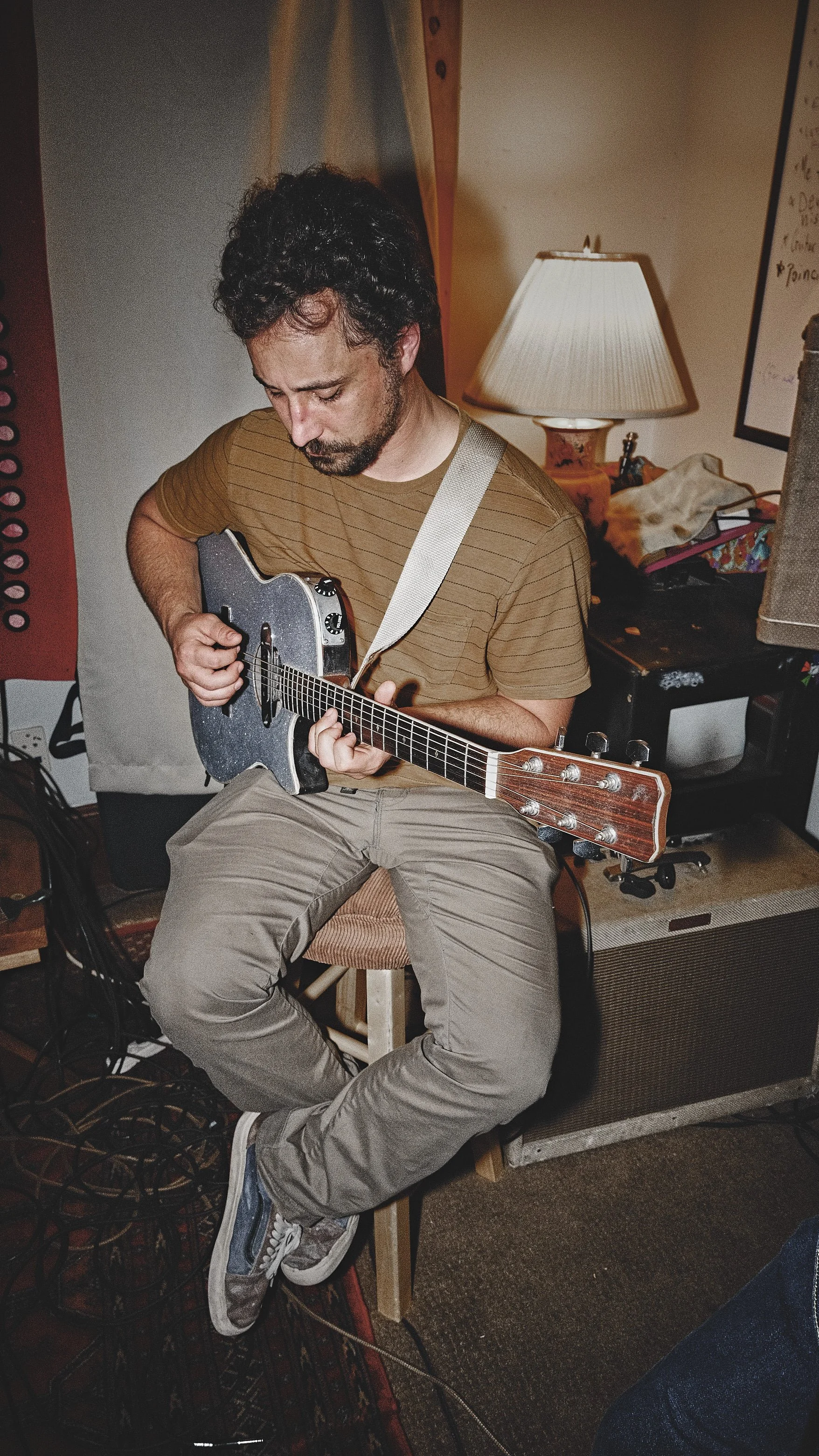 A man with dark curly hair and a beard sits on a stool, playing an electric guitar in a cozy room with warm lighting. He is wearing a striped brown t-shirt and beige pants.