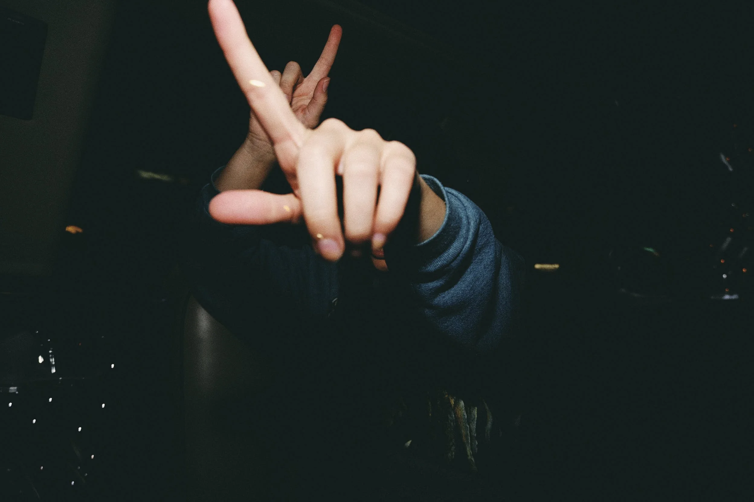 Person making a peace sign with their hand in front of the camera, wearing a dark jacket, with a dark background.
