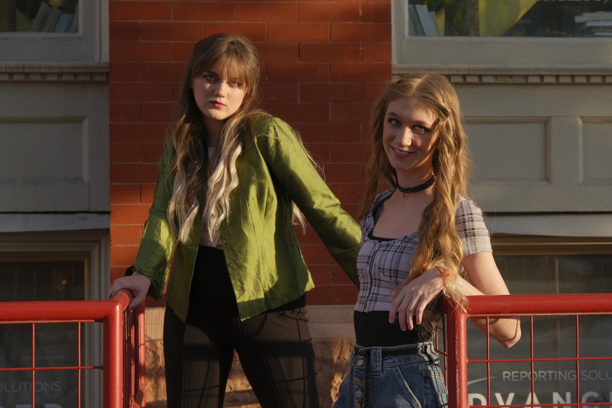 Two young women standing outside near a red railing in front of a brick building, one with long blond hair and a green jacket, the other with long wavy red hair and a plaid top, smiling at the camera during golden hour.