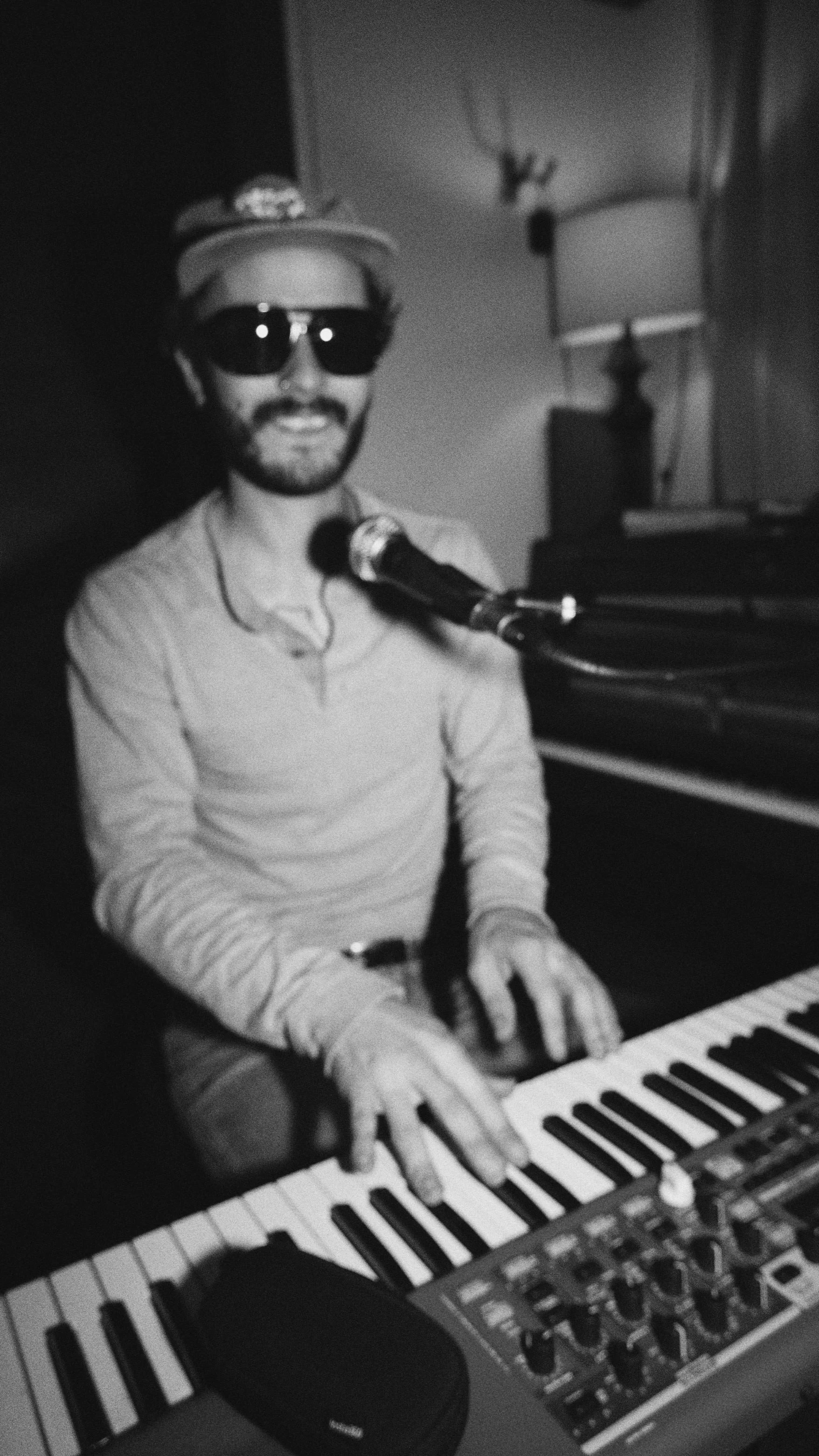 A man wearing sunglasses and a hat, sitting at a piano, playing it, with a microphone positioned near his mouth.