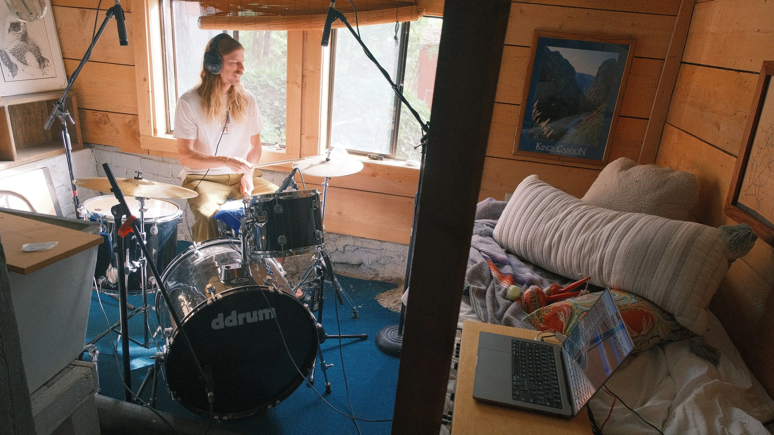 A woman playing drums in a cozy, rustic room with wooden walls and large windows. There is a bed and a laptop visible in the room.