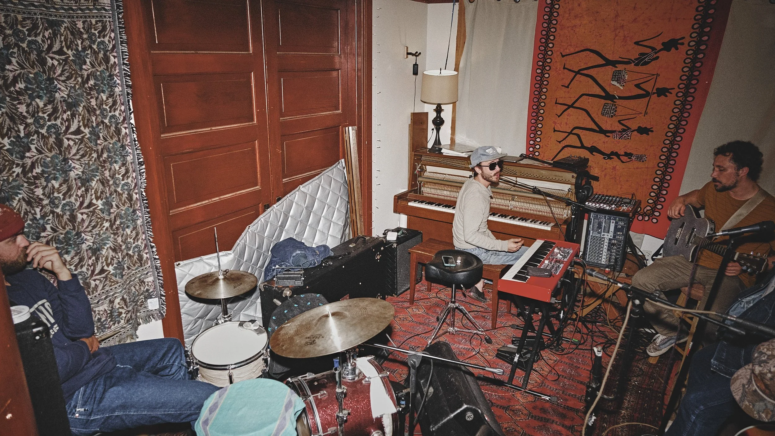 A band of three musicians in a cozy, decorated room, with acoustic and electronic instruments. One person plays guitar, another is seated at a keyboard with a microphone, and a third is on drums. The room has patterned rugs and wall hangings.