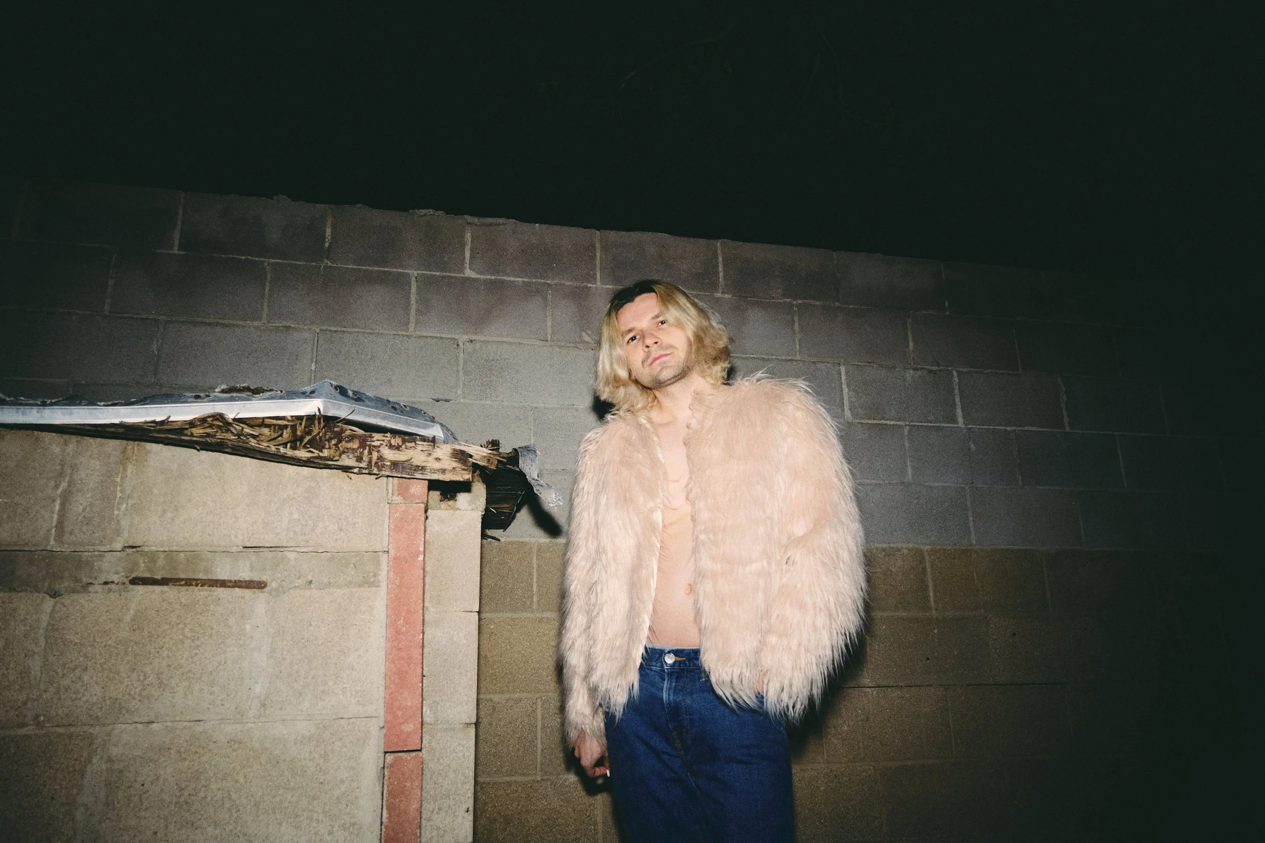 A person with blonde hair wearing a fluffy pink jacket and blue jeans, standing against a concrete wall in a dimly lit environment.