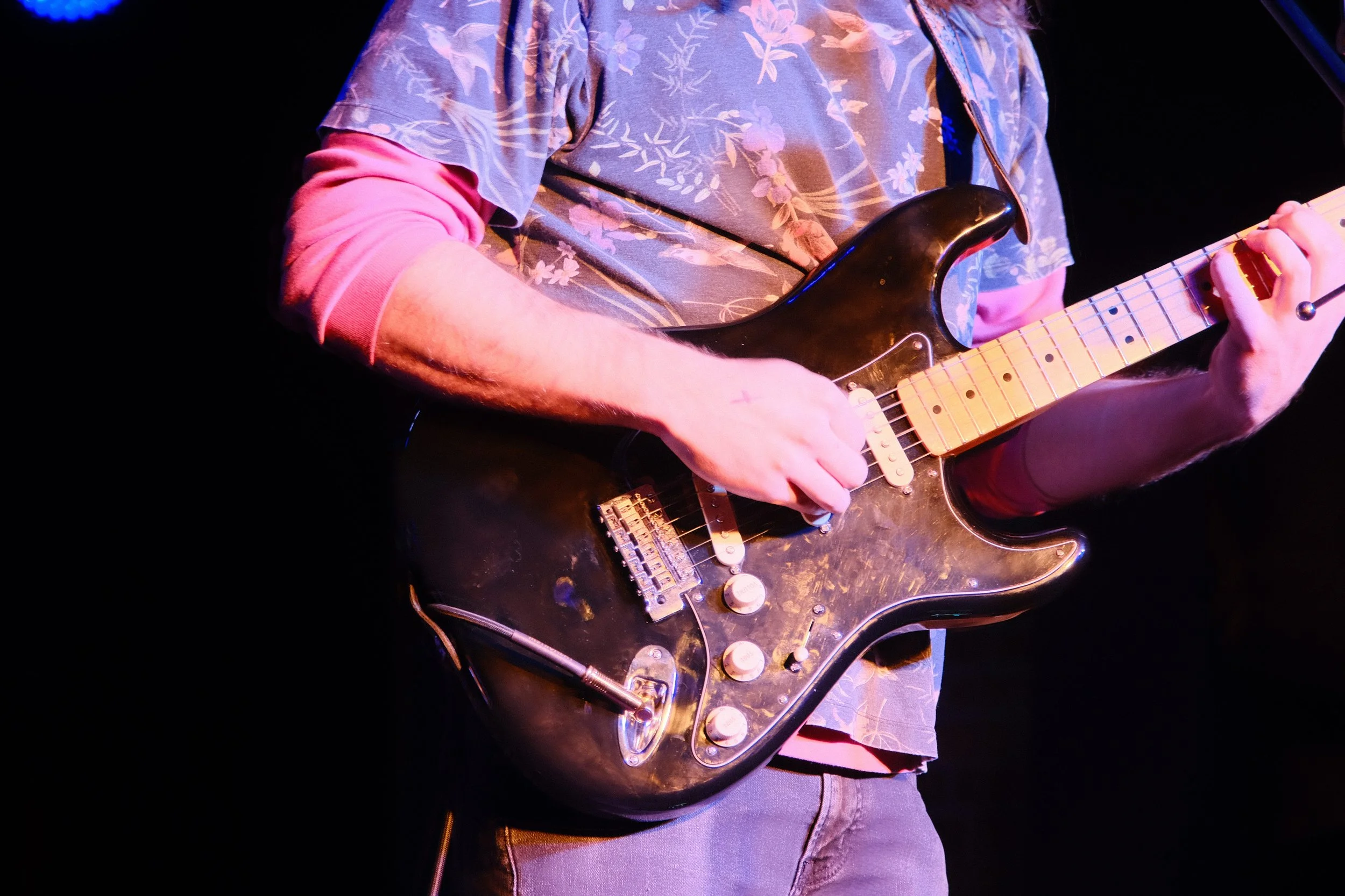 Person playing a black electric guitar on stage, wearing a floral shirt with pink sleeves.