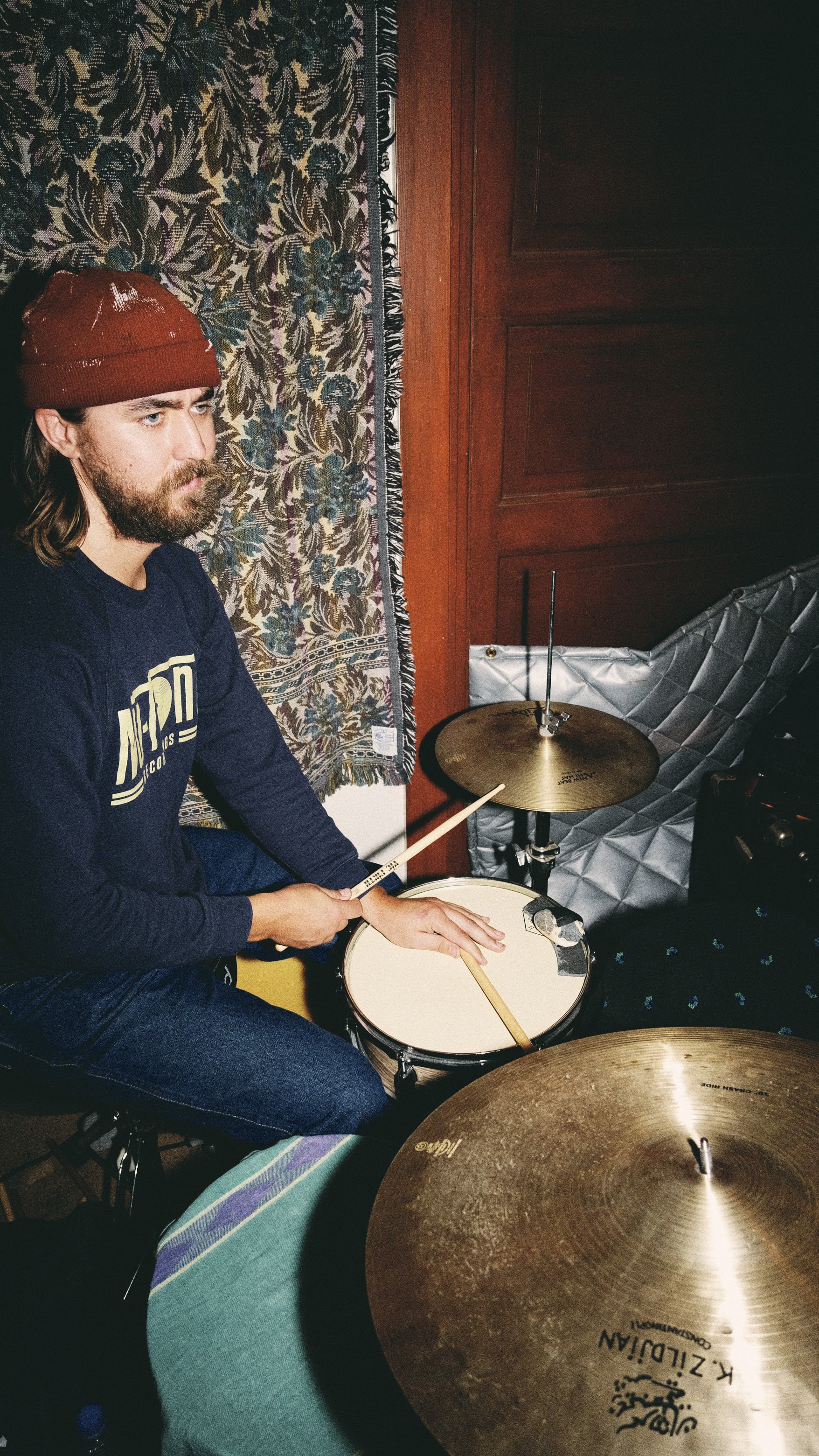 A man with long hair, beard, wearing a red beanie and a dark blue shirt, sitting behind a drum set. He holds a drumstick in his right hand and rests his left hand on a snare drum. Behind him, there is a patterned tapestry and a wooden wall.