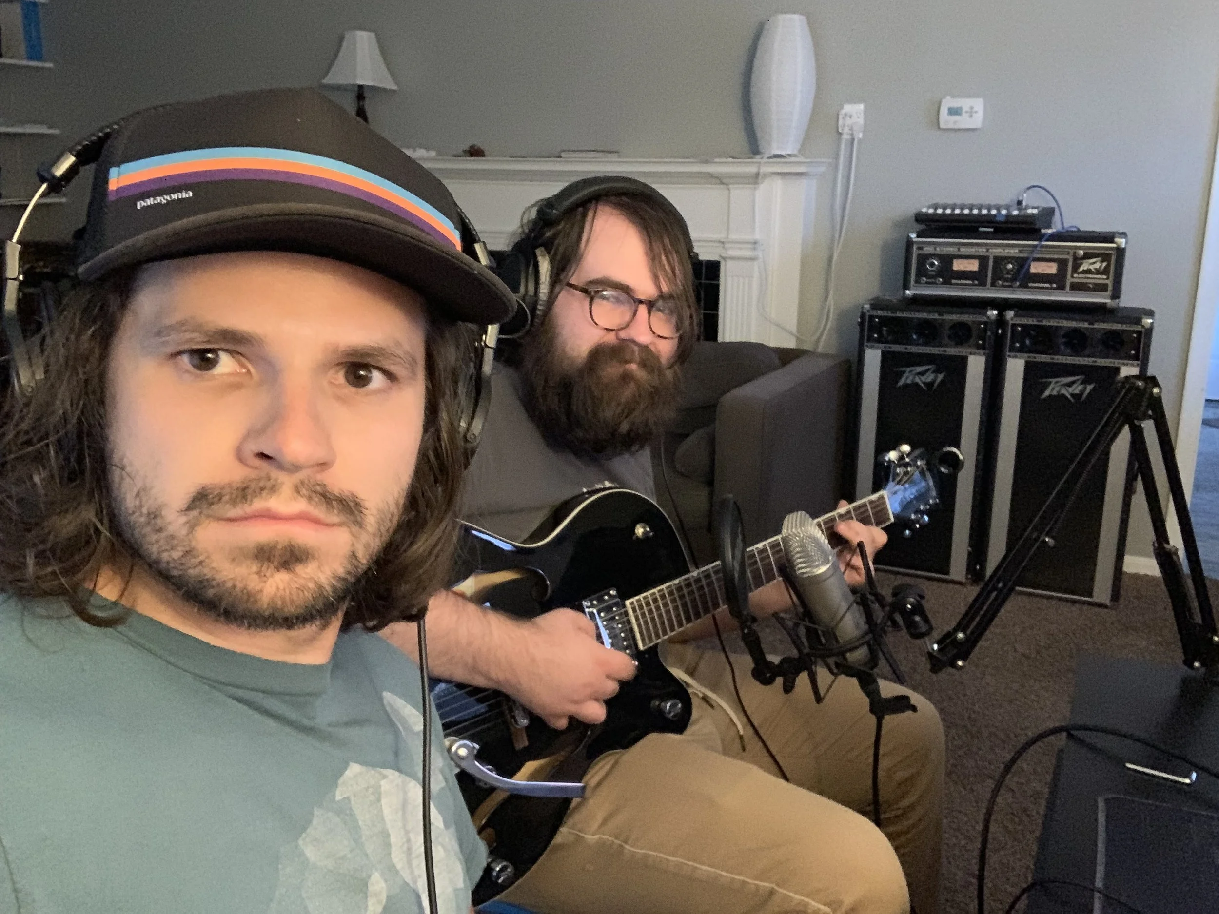 Two men recording music in a home studio: one in the foreground wearing a Patagonia cap and headphones, the other in the background playing an electric guitar, with audio equipment in the room