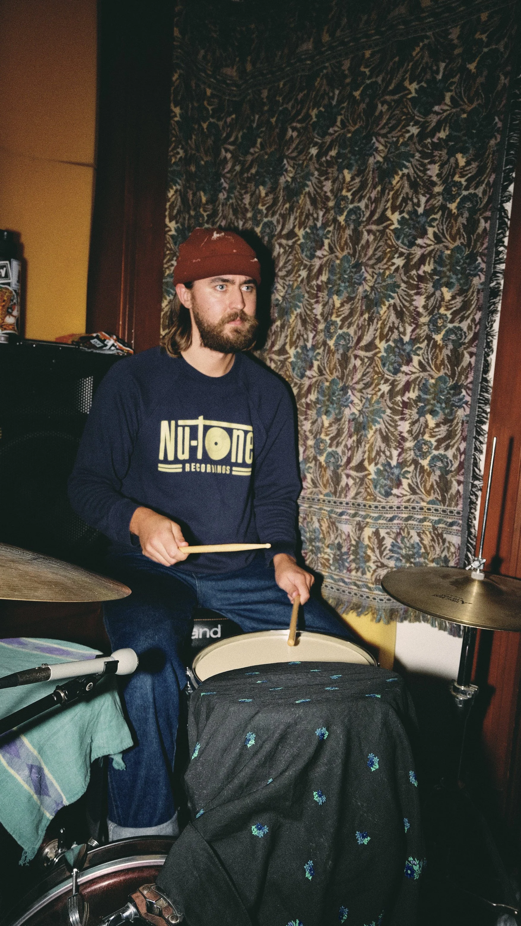 A man with long hair, a beard, wearing a red beanie and a navy blue sweatshirt with 'Nu-Tone Recordings' printed on it, is sitting on a chair and playing a drum set in a room with a decorative tapestry on the wall.