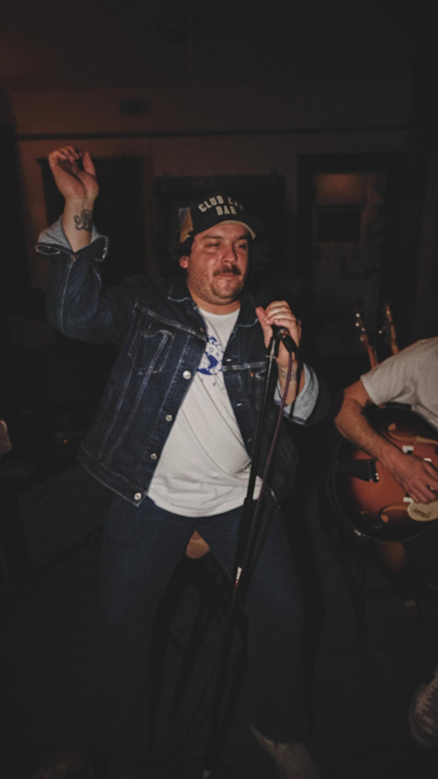 A man singing into a microphone, wearing a denim jacket and a 'Club House' cap, with another person playing an acoustic guitar beside him.