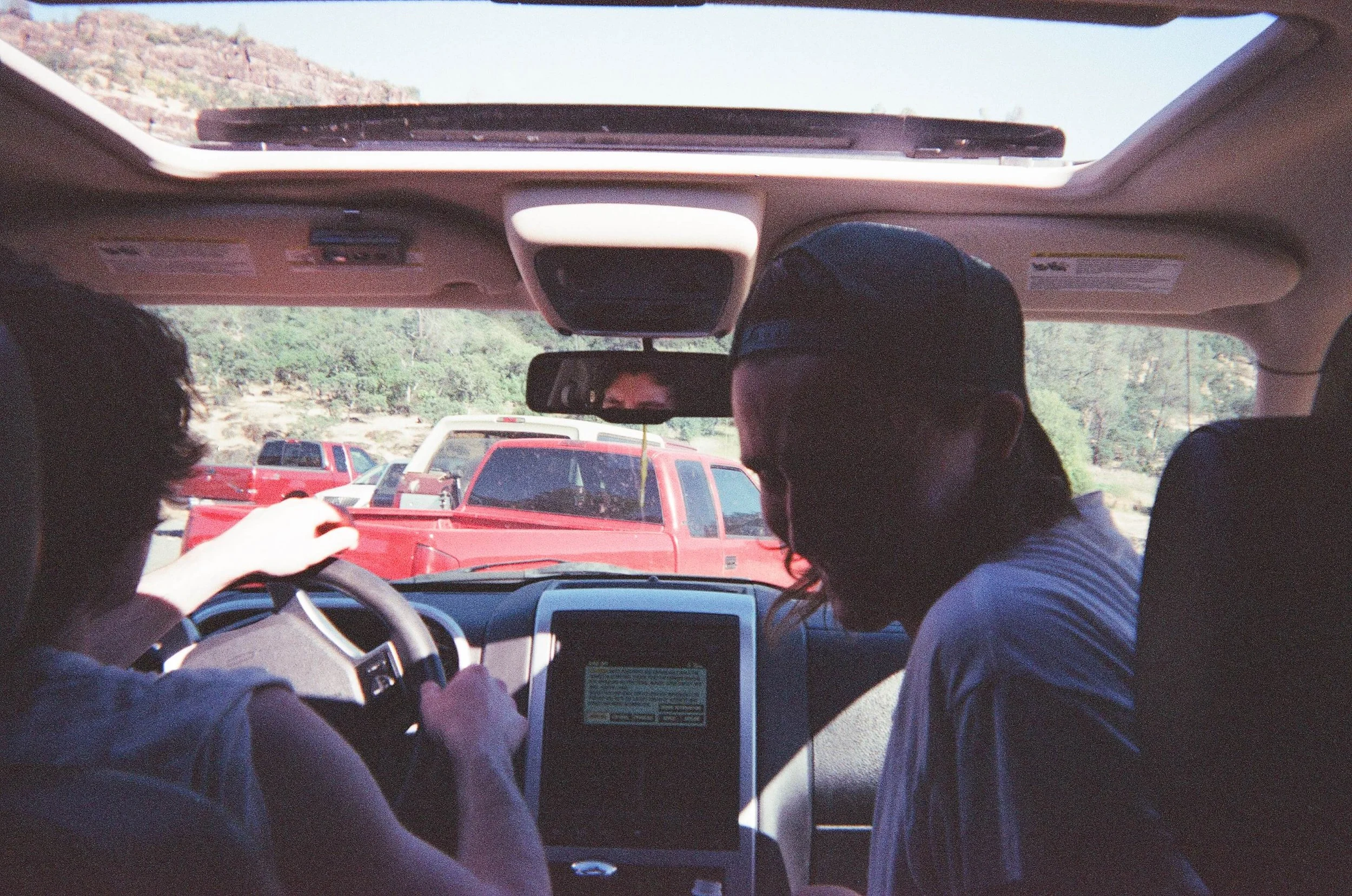 People in a vehicle with one person driving and another person sitting in the passenger seat, visible through the windshield with a view of parked trucks and trees outside.
