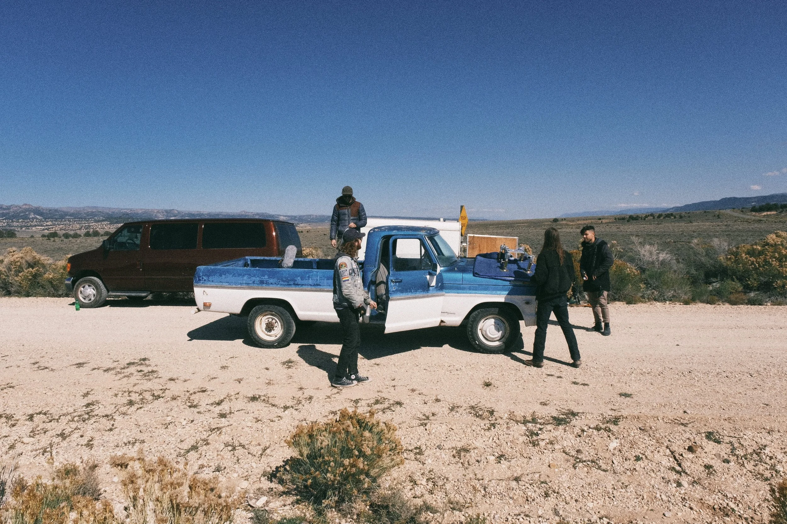 A group of people standing near a blue and white pickup truck in a desert landscape on a sunny day, with hills and sparse bushes in the background.