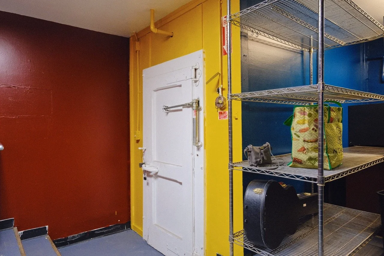 Steel storage shelves with a black case, green tote bag, and a small gray bag in a room with yellow, blue, and maroon walls.