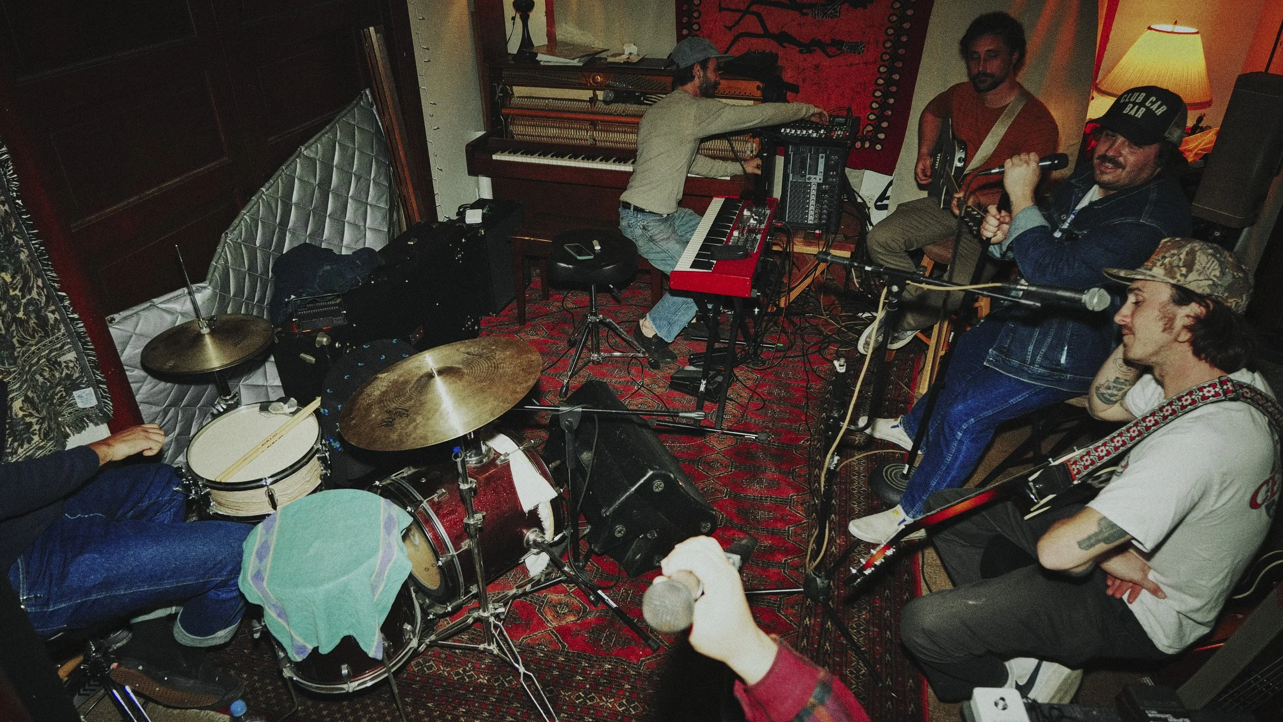 Five band members in a recording studio, with one playing drums, one on keyboard, two on guitar, and one singing into a microphone.