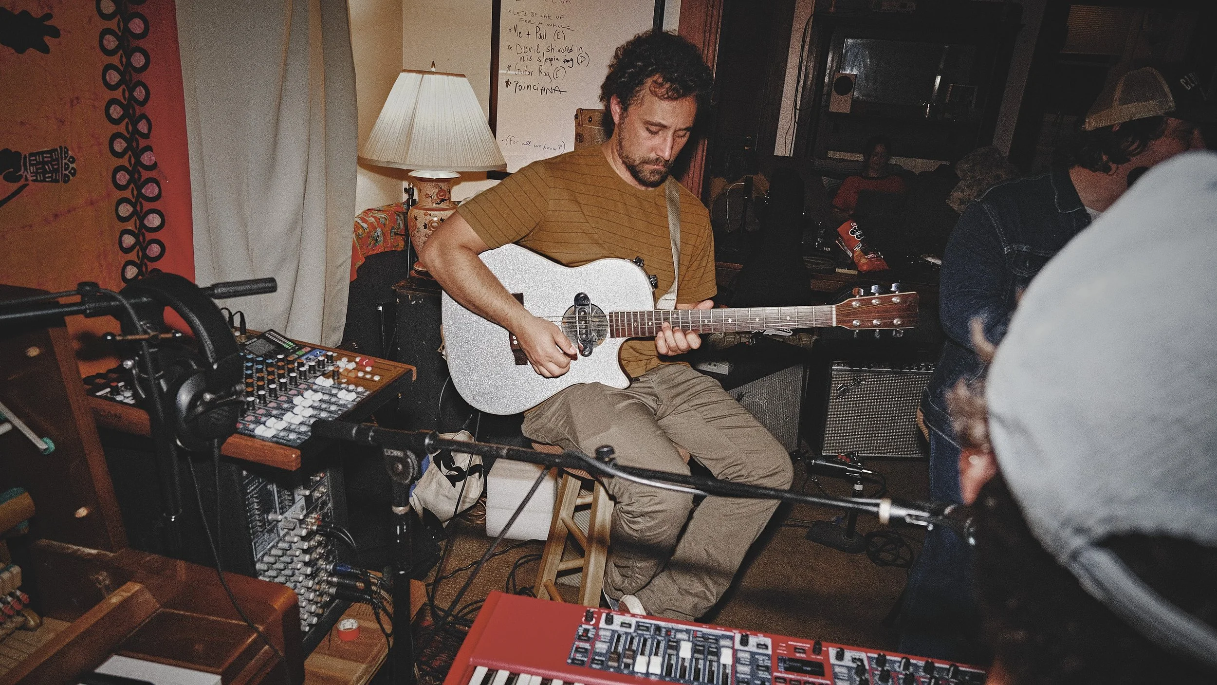 A man playing an acoustic guitar in a dimly lit room filled with music equipment and people in the background.