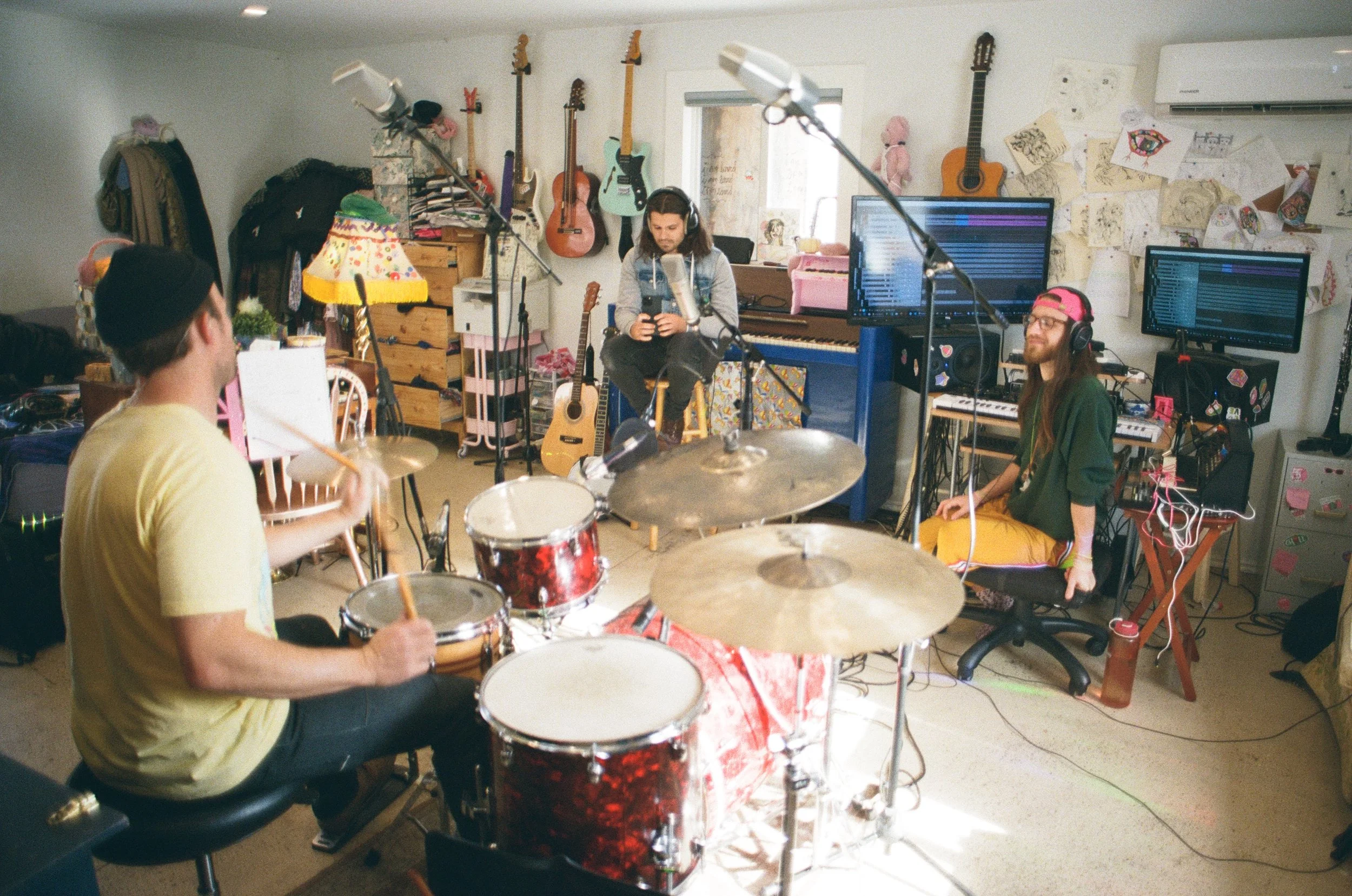 A band of three young musicians rehearsing in a cluttered music studio filled with guitars, computers, and artwork