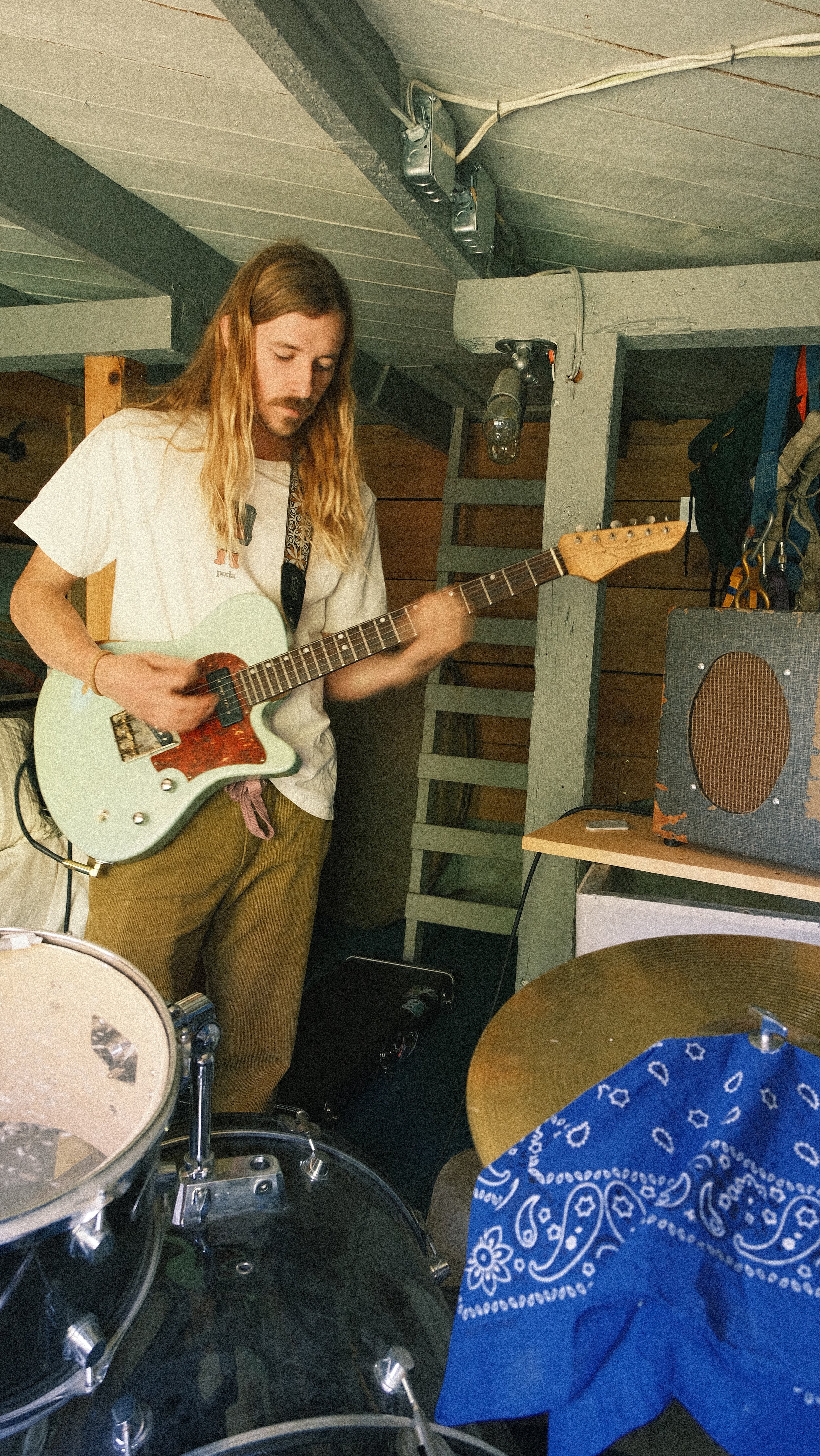A man playing an electric guitar in a music studio or practice space with various instruments and equipment around him.