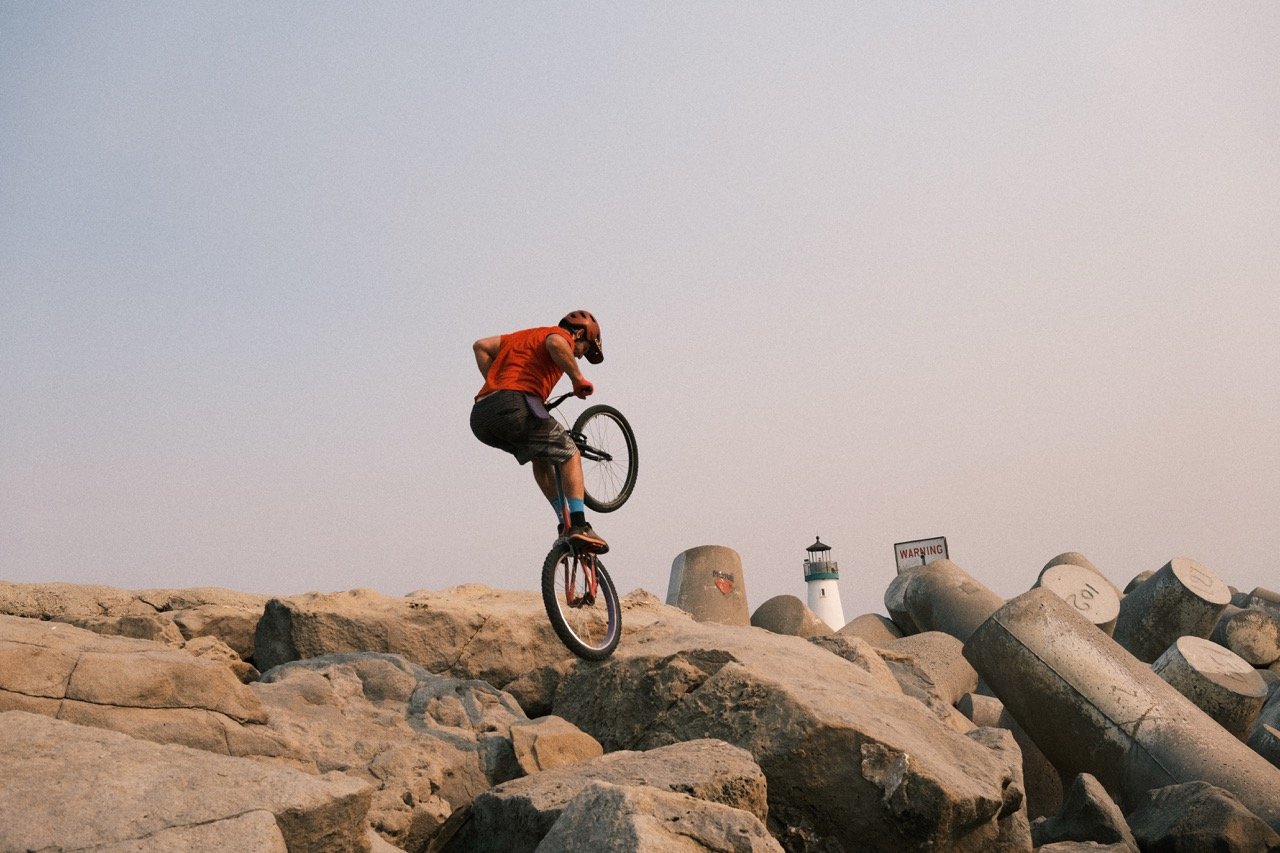 A person wearing an orange shirt, helmet, and shorts riding a mountain bike on large rocks near a coastline with a small lighthouse in the background under a pale sky.