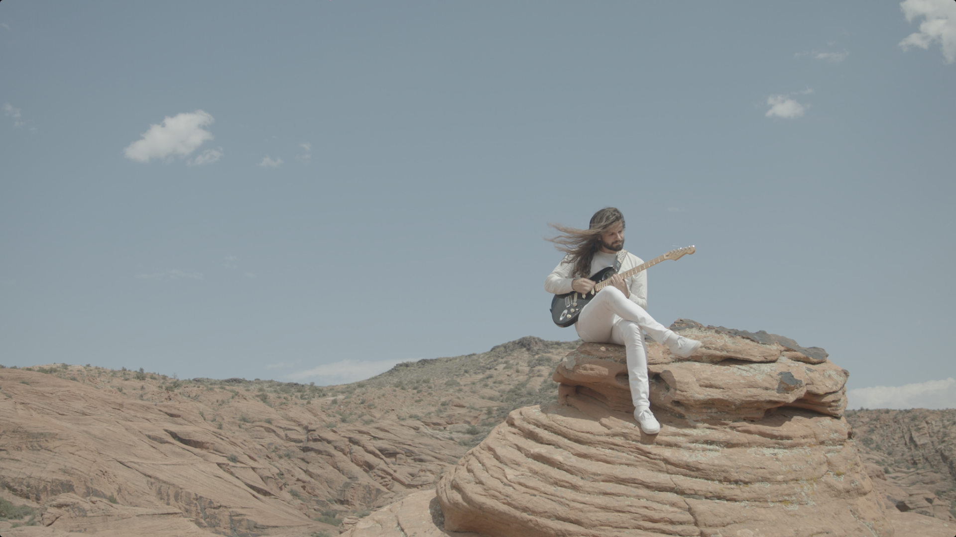 A man with long hair, wearing white pants and a white shirt, sits on a large rock formation playing an electric guitar in a desert landscape with a blue sky and a few clouds.