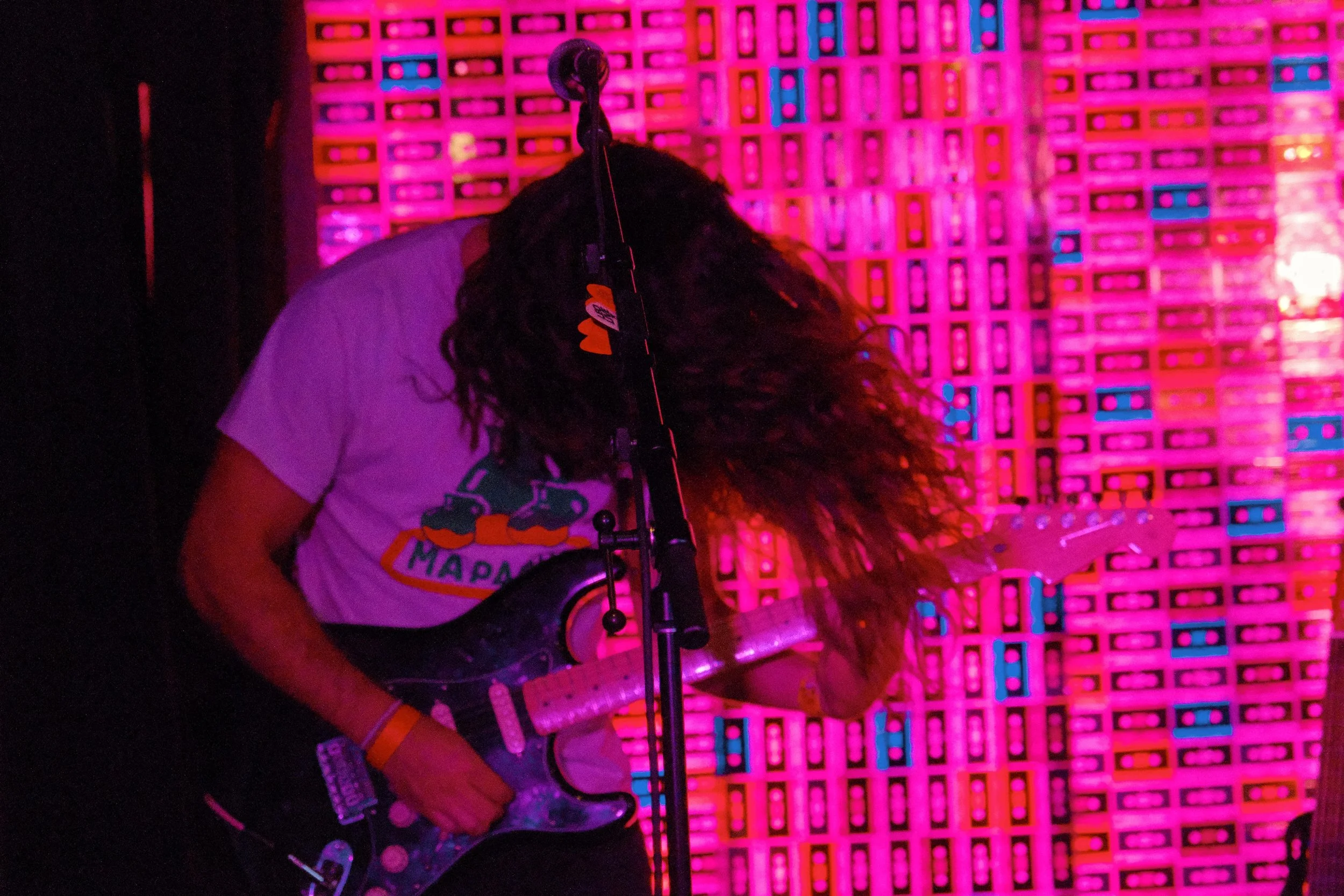 A musician with long curly hair playing an electric guitar on stage, in front of a neon pink and blue background, with a microphone stand nearby.