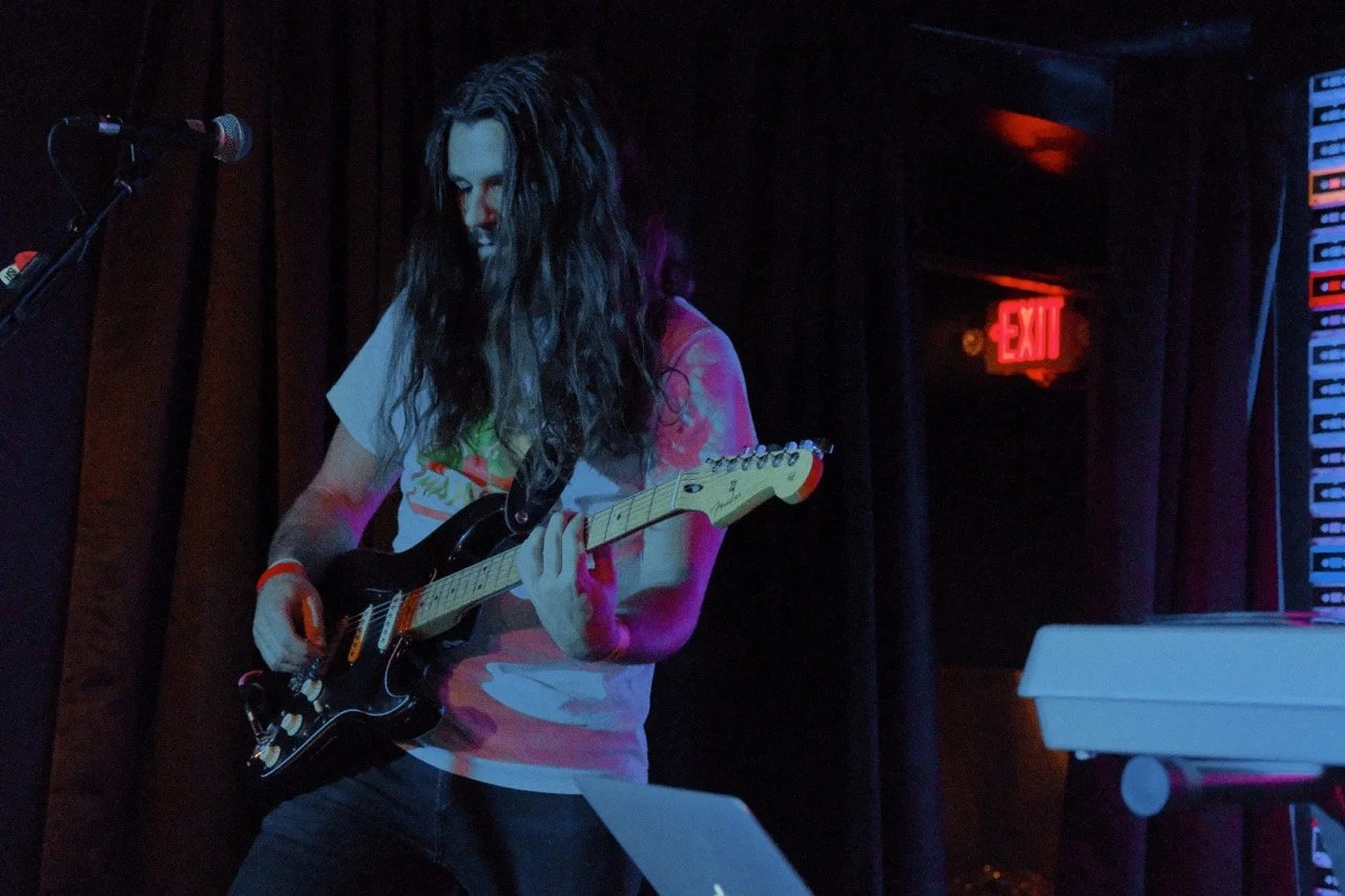 A person with long dark hair playing an electric guitar on stage with a dark background and a red EXIT sign.
