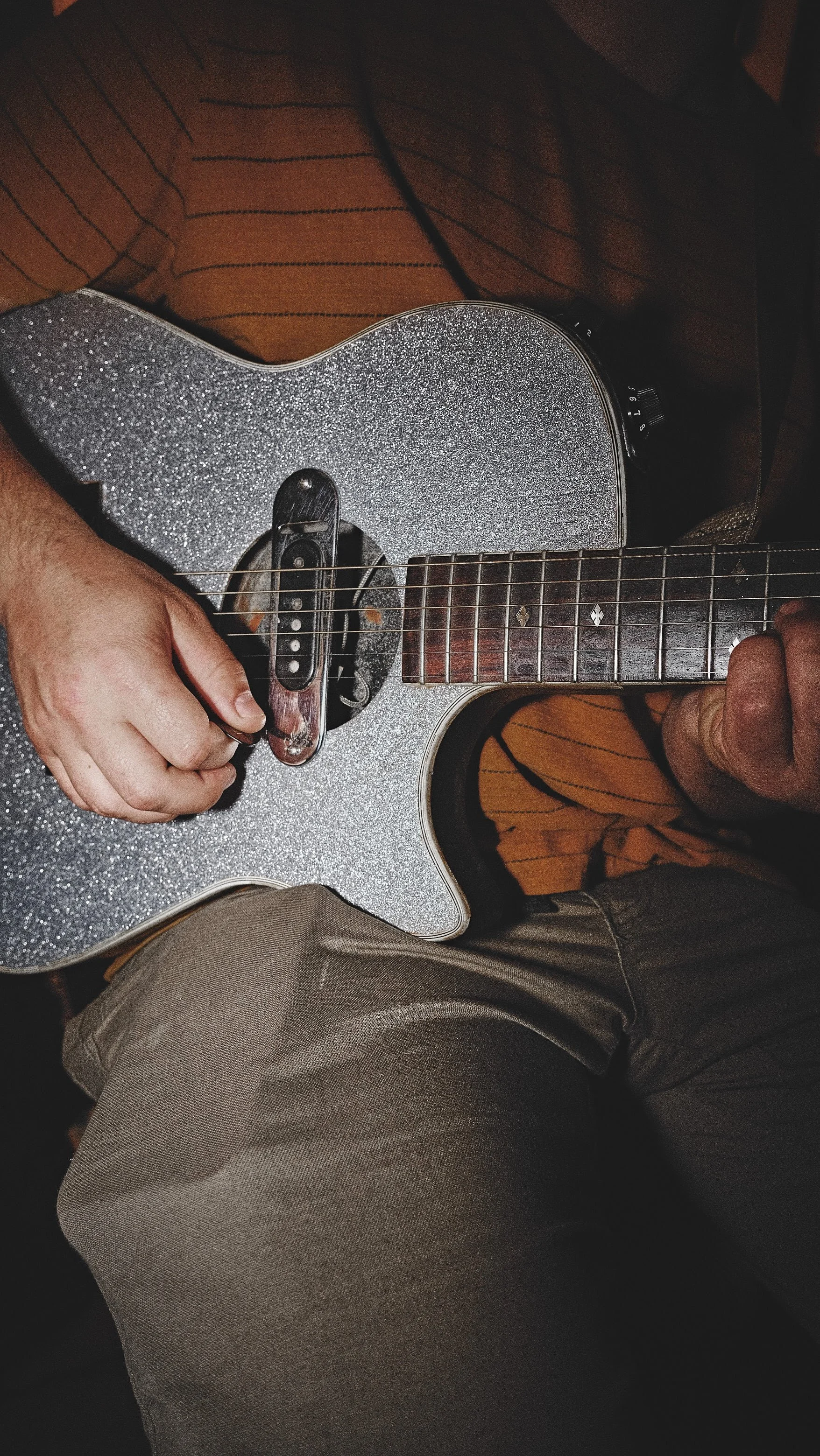 A person playing an acoustic guitar with a glittery silver body, wearing beige pants and a striped orange and black shirt.
