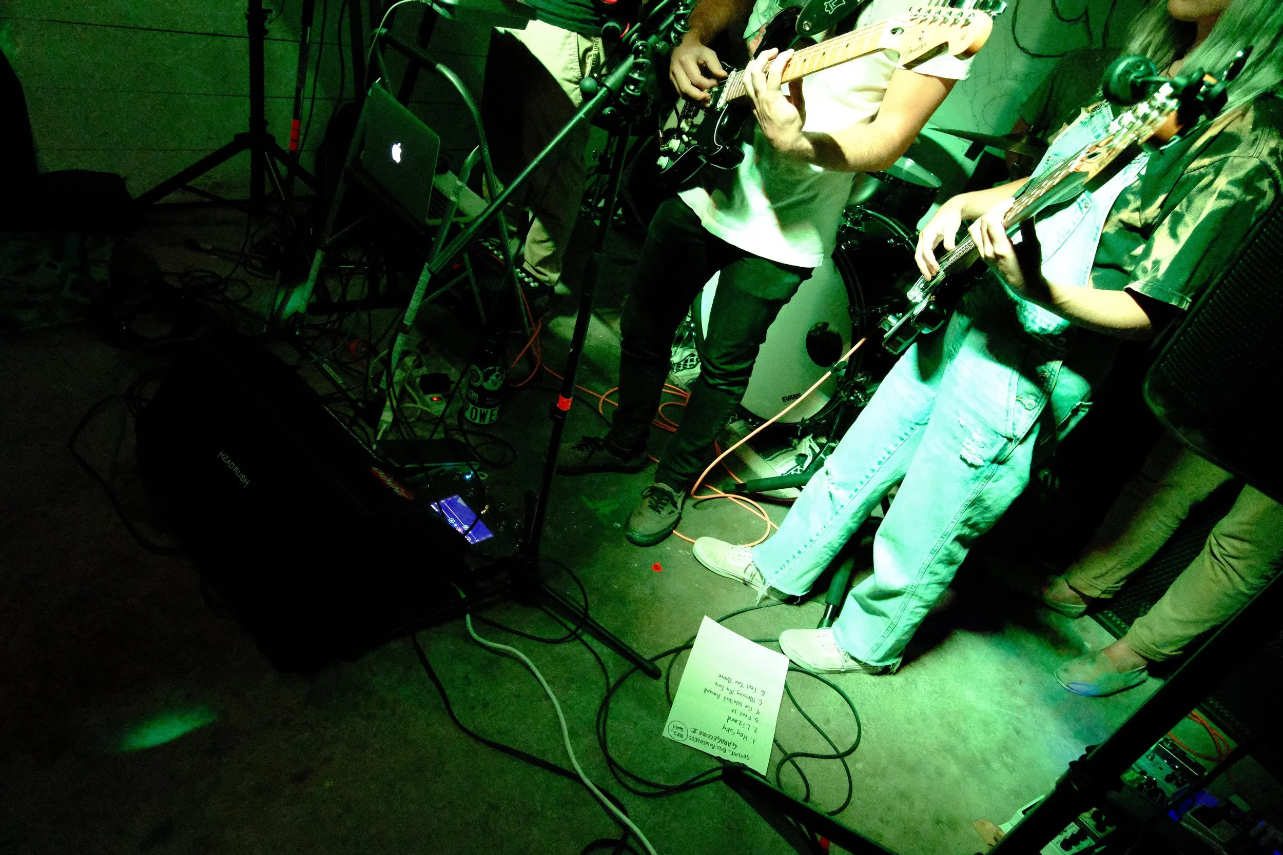 People playing electric guitars on stage with music equipment and a setlist on the floor, lit with green stage lights.