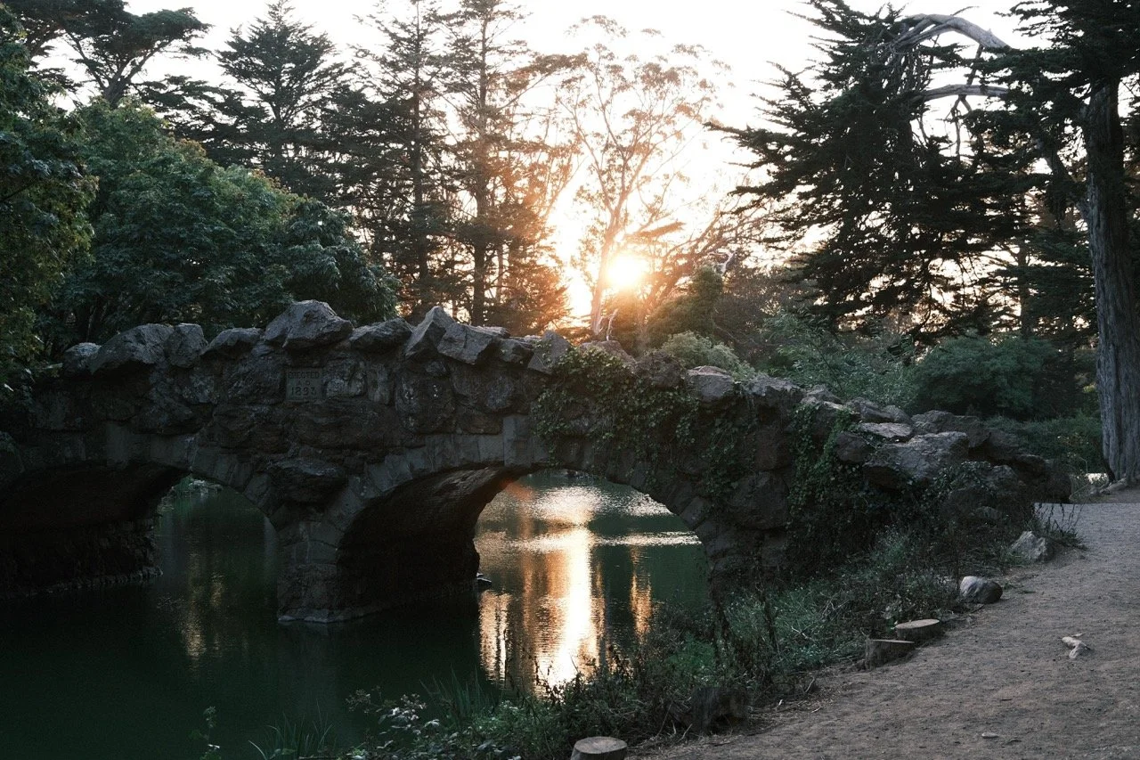 A stone bridge over a calm body of water with trees and a sunset in the background.