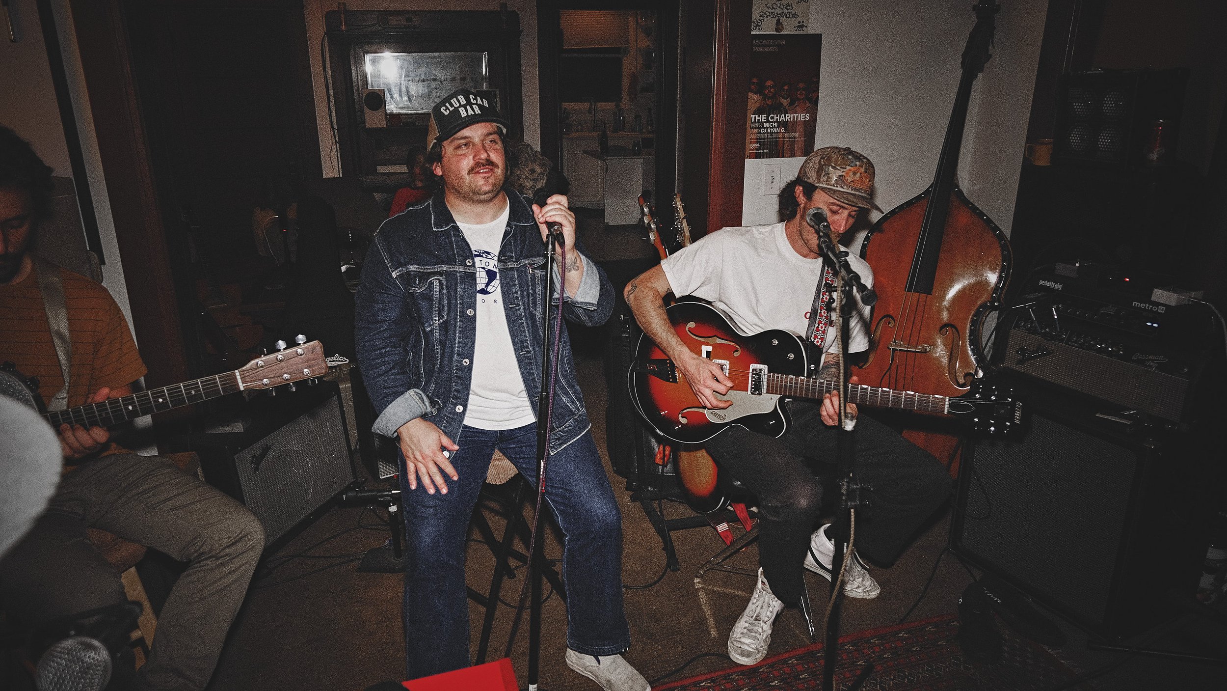 A band performing live in an indoor venue with three musicians: one playing an acoustic guitar, one singing into a microphone, and another playing an electric guitar. There is a double bass behind them and various music equipment around.
