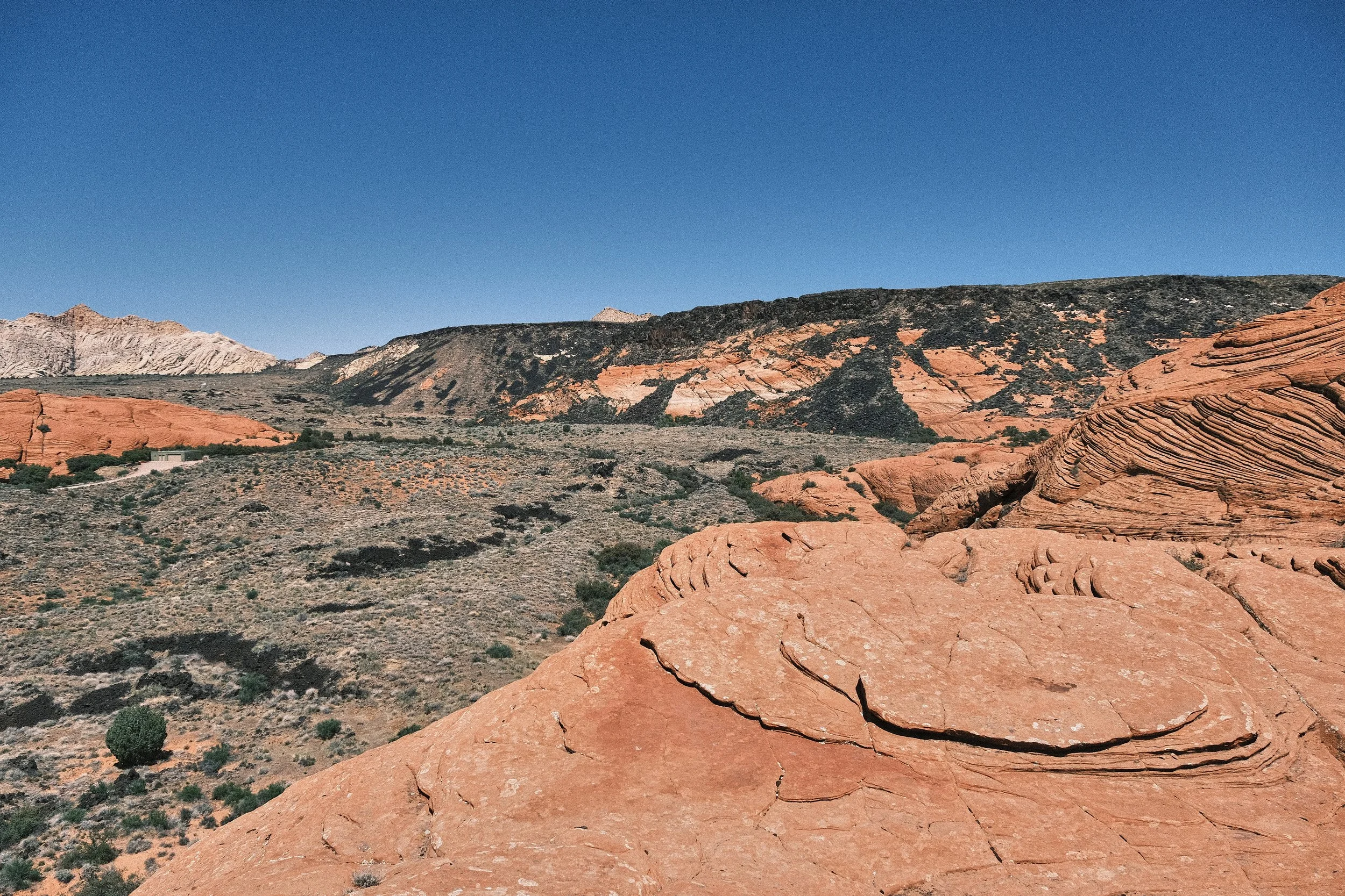 Desert landscape with large reddish rock formations, sparse vegetation, and a distant mountain range under a clear blue sky.