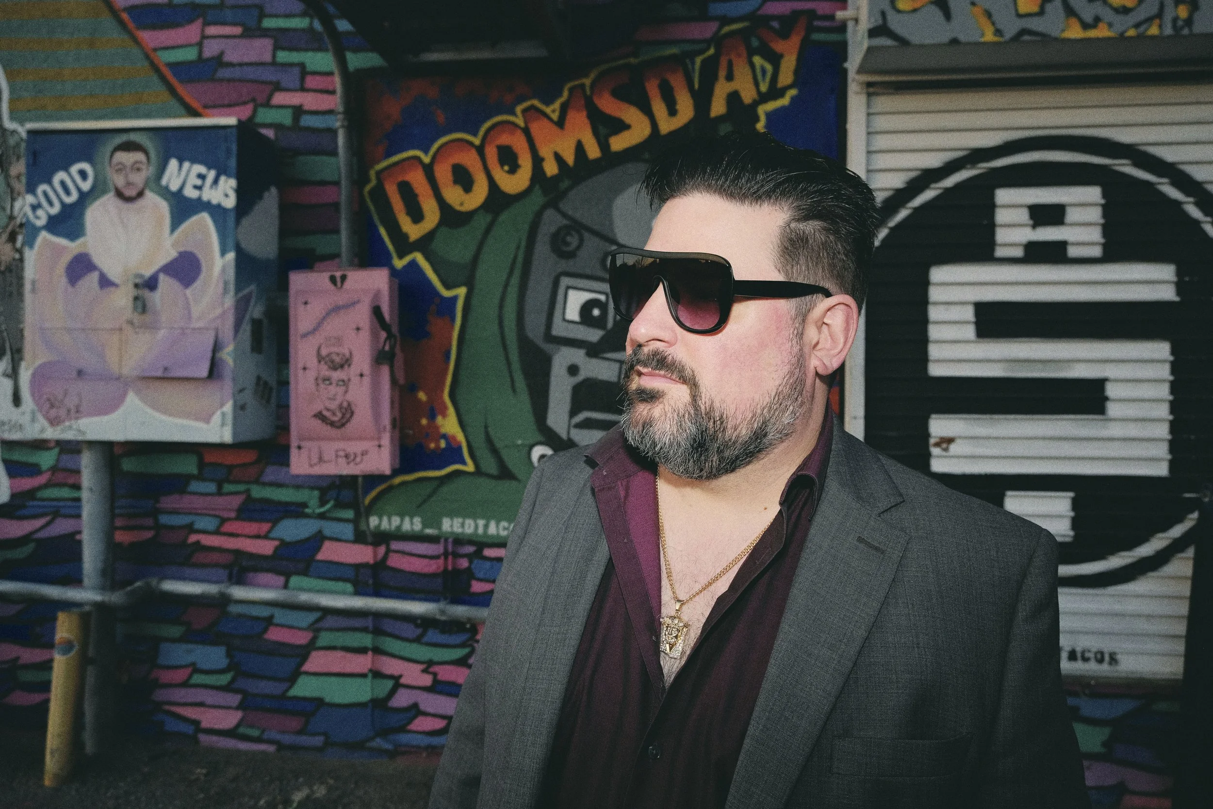 A man with dark hair, beard, and sunglasses wearing a gray suit and a maroon shirt with an open collar, standing in front of colorful graffiti art that includes the words 'DOOMSDAY' and various cartoon-like images.