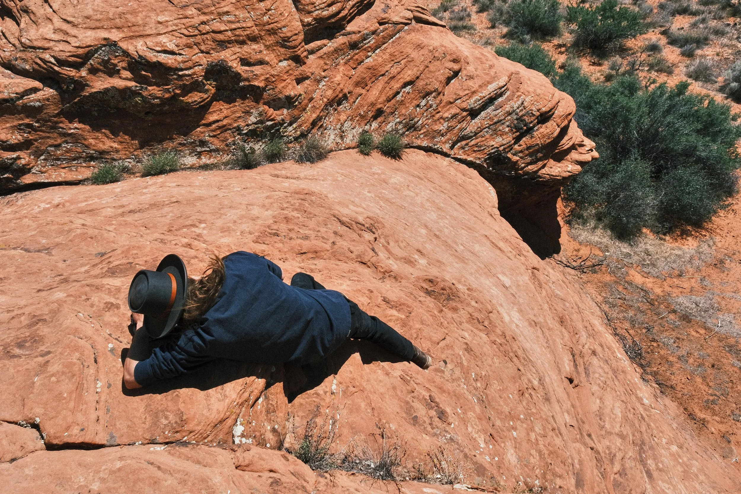 Person climbing a steep, reddish rock formation in a desert landscape with scattered green bushes.