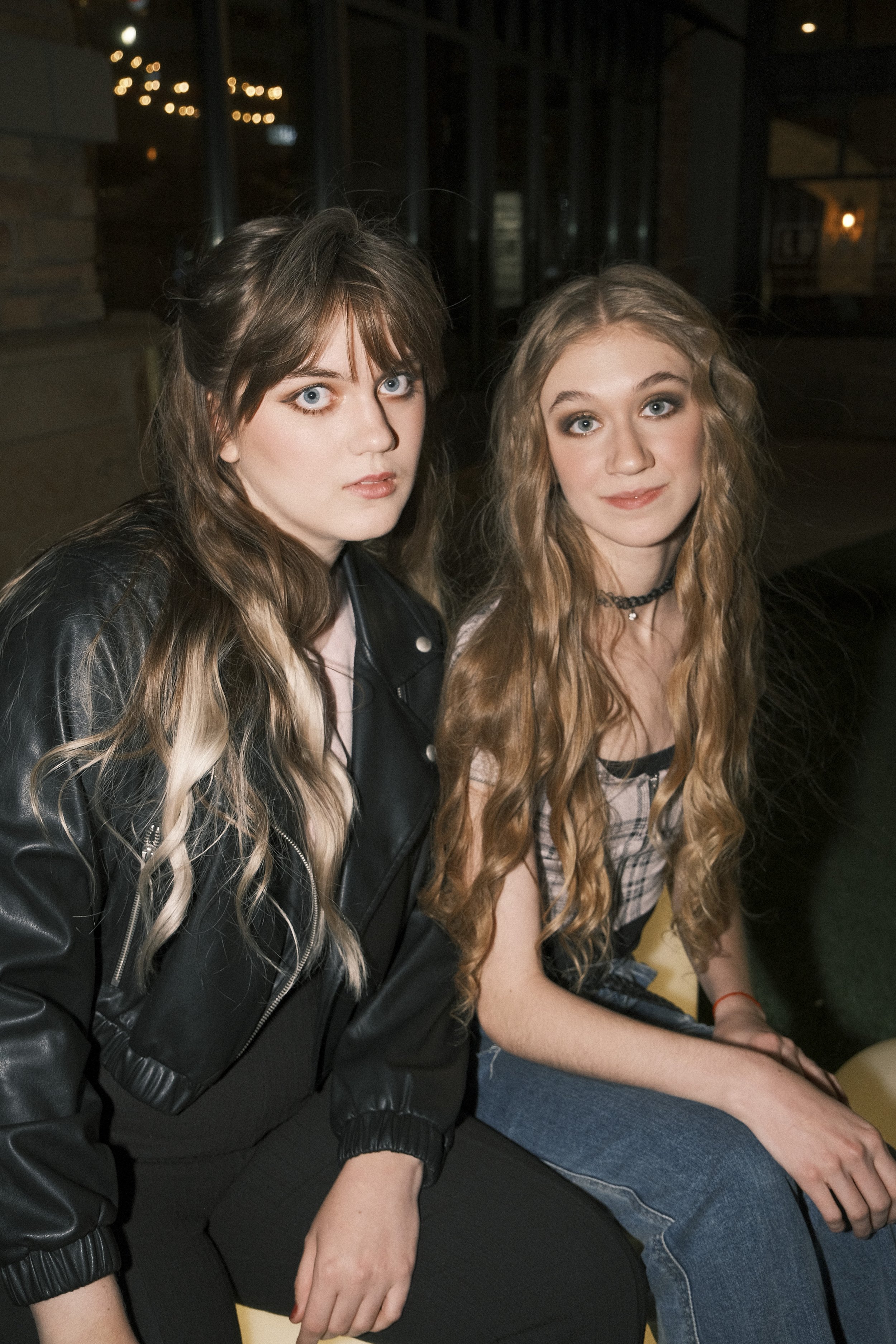 Two young women sitting indoors at night, one in a black leather jacket with blond hair and blue eyes, the other with long red hair and a plaid shirt, smiling at the camera.
