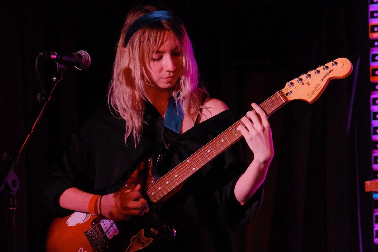 A woman with blonde hair wearing a black headband and black top, playing an electric guitar on stage with pink and purple lighting.