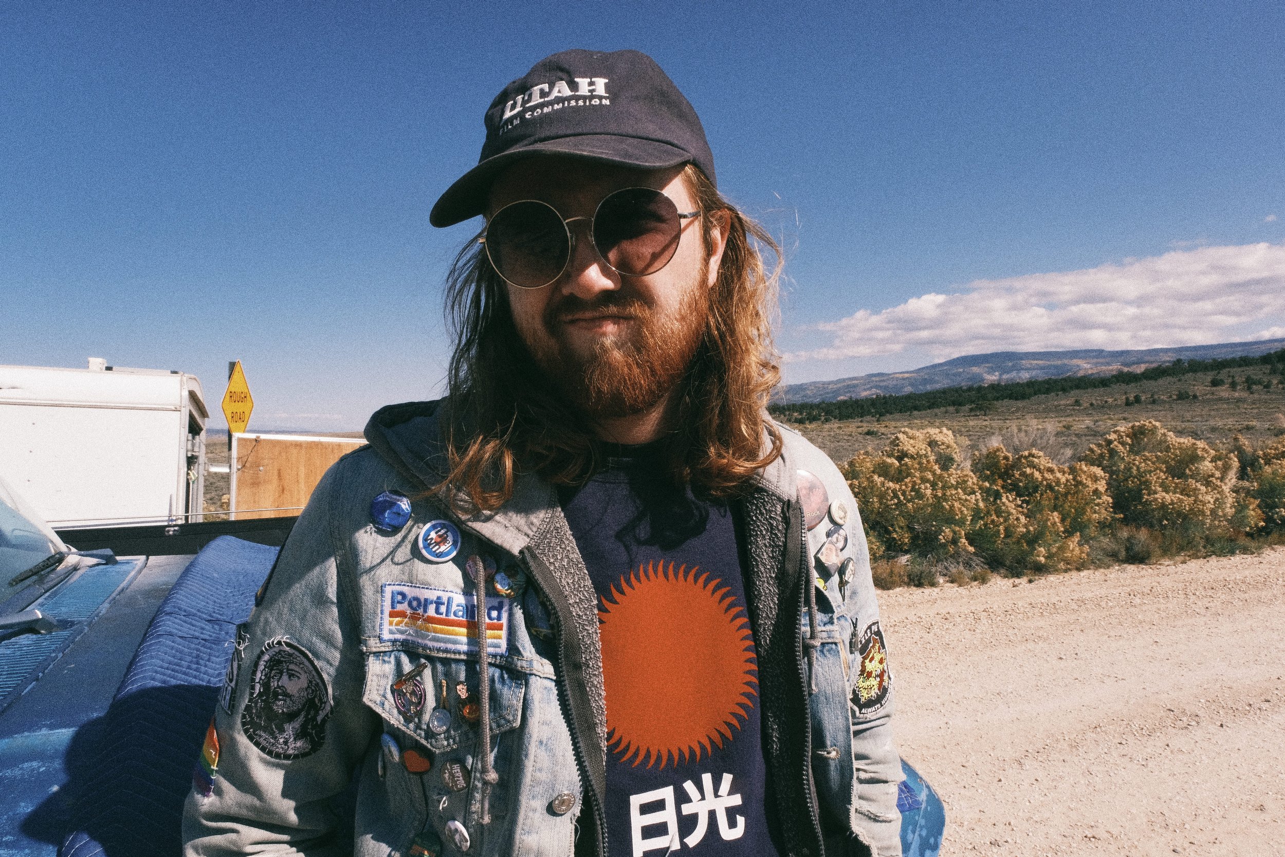 A man with long hair and beard wearing a dark cap, sunglasses, a denim jacket with patches, and a black T-shirt with an orange sun design and Japanese characters, standing outdoors with a desert landscape, bushes, mountains, and a blue sky in the bac